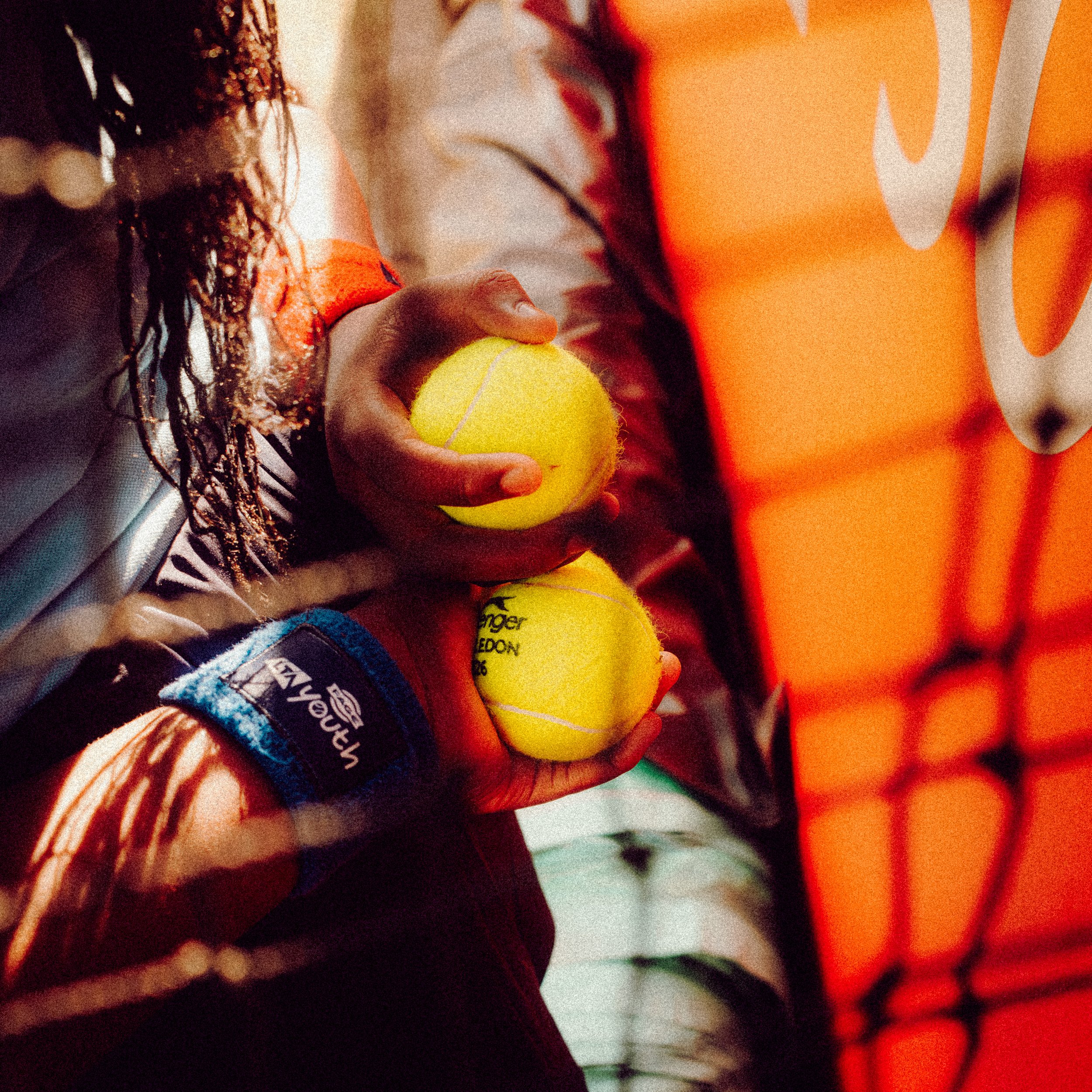 Close-up of person’s hand holding three yellow tennis balls, with one ball facing outward and showing text, against a background of a bright orange wall with white and red design.