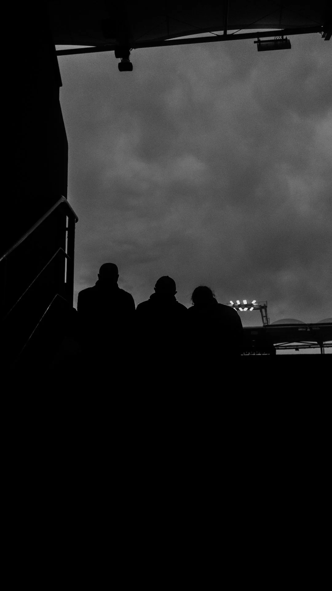 Silhouettes of three people standing outdoors with cloudy sky in the background, partially visible metal structures and stairs on the left.