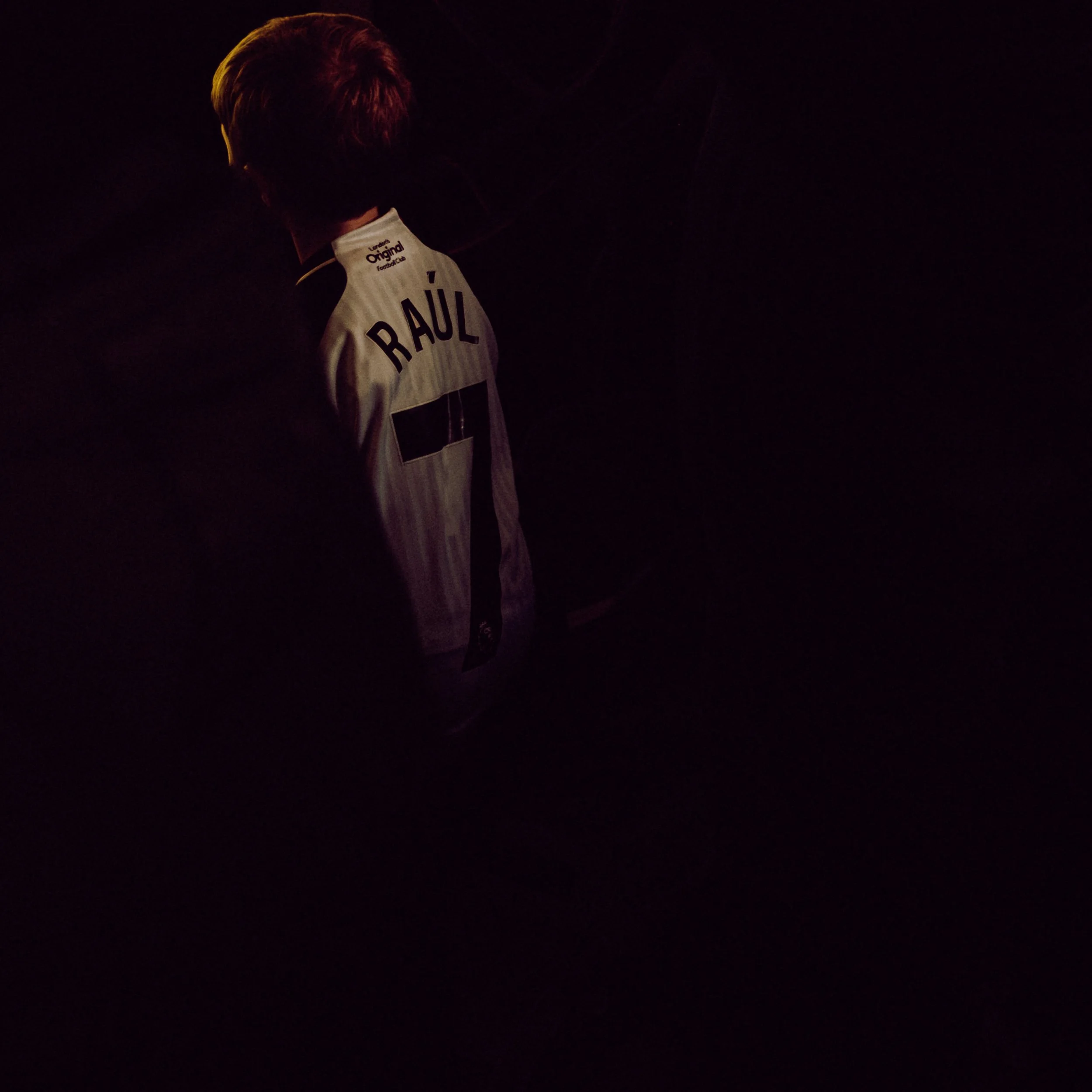 A person is wearing a white and purple soccer jersey with the name 'RÁUL' printed on the back, standing in a dark environment with faint reddish lighting on their hair.