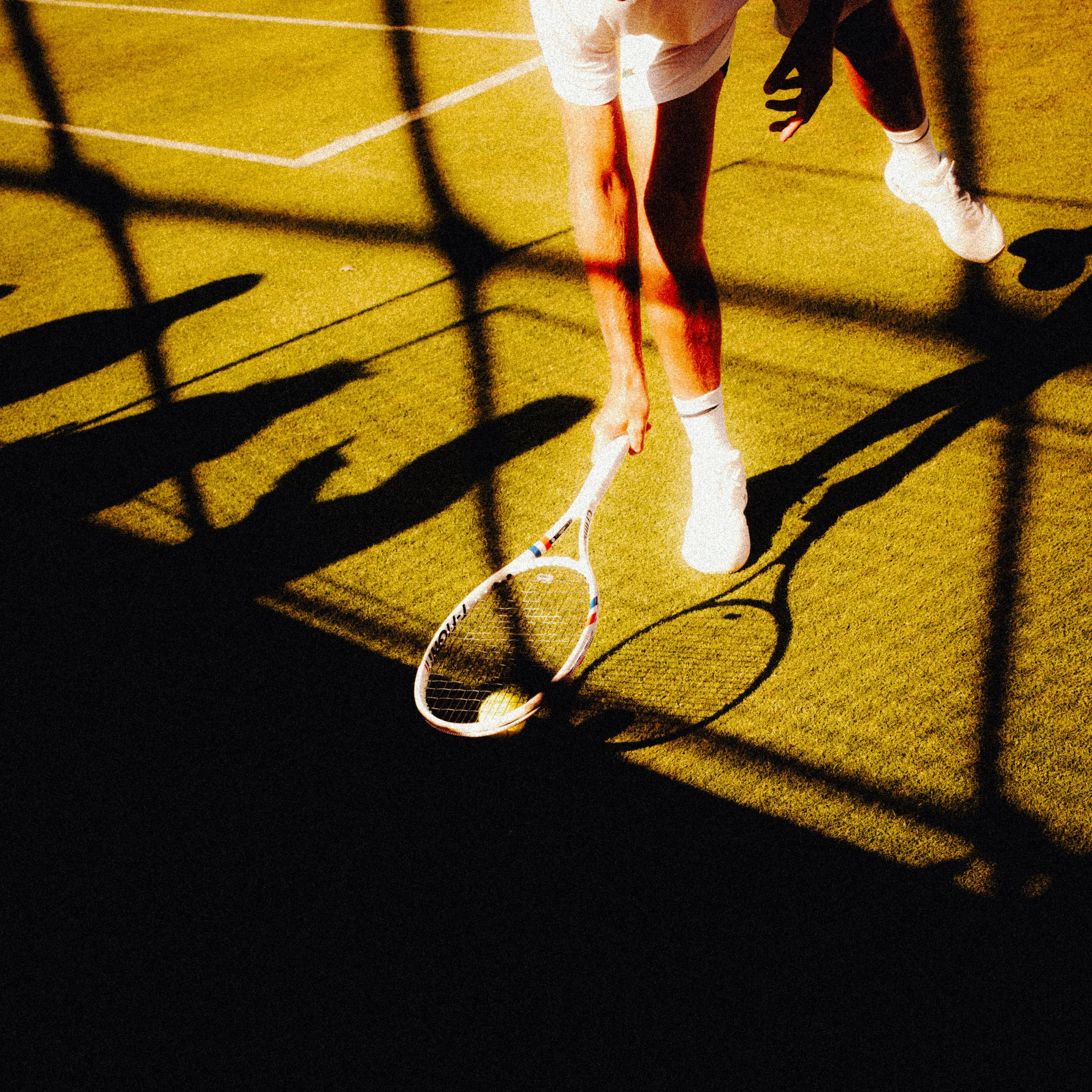 A player on a tennis court, kneeling with one knee on the ground, wearing white athletic clothing and holding a tennis racket, with shadows cast on the court.