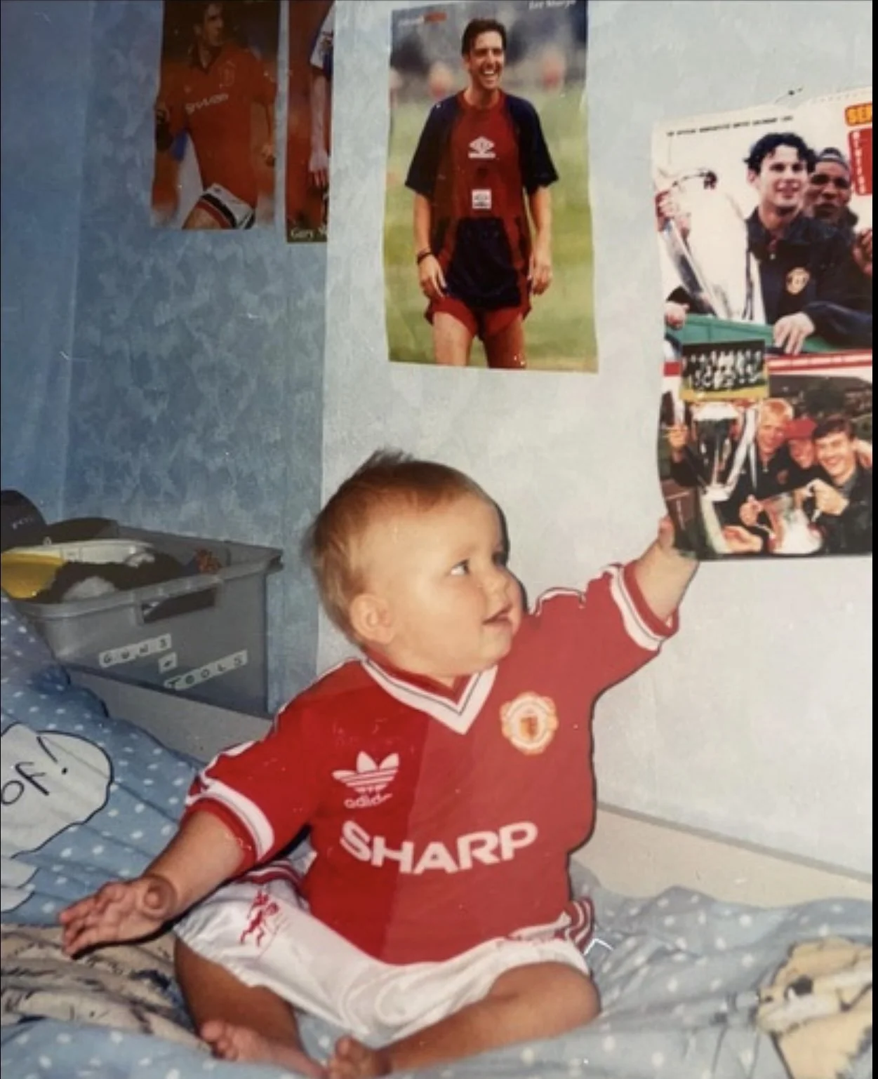 A young boy with red hair and light skin wearing a red Manchester United jersey, sitting on a bed, and reaching out to touch a poster on the wall. The poster features a man in a sports uniform smiling outdoors. The boy is looking at the poster with curiosity.