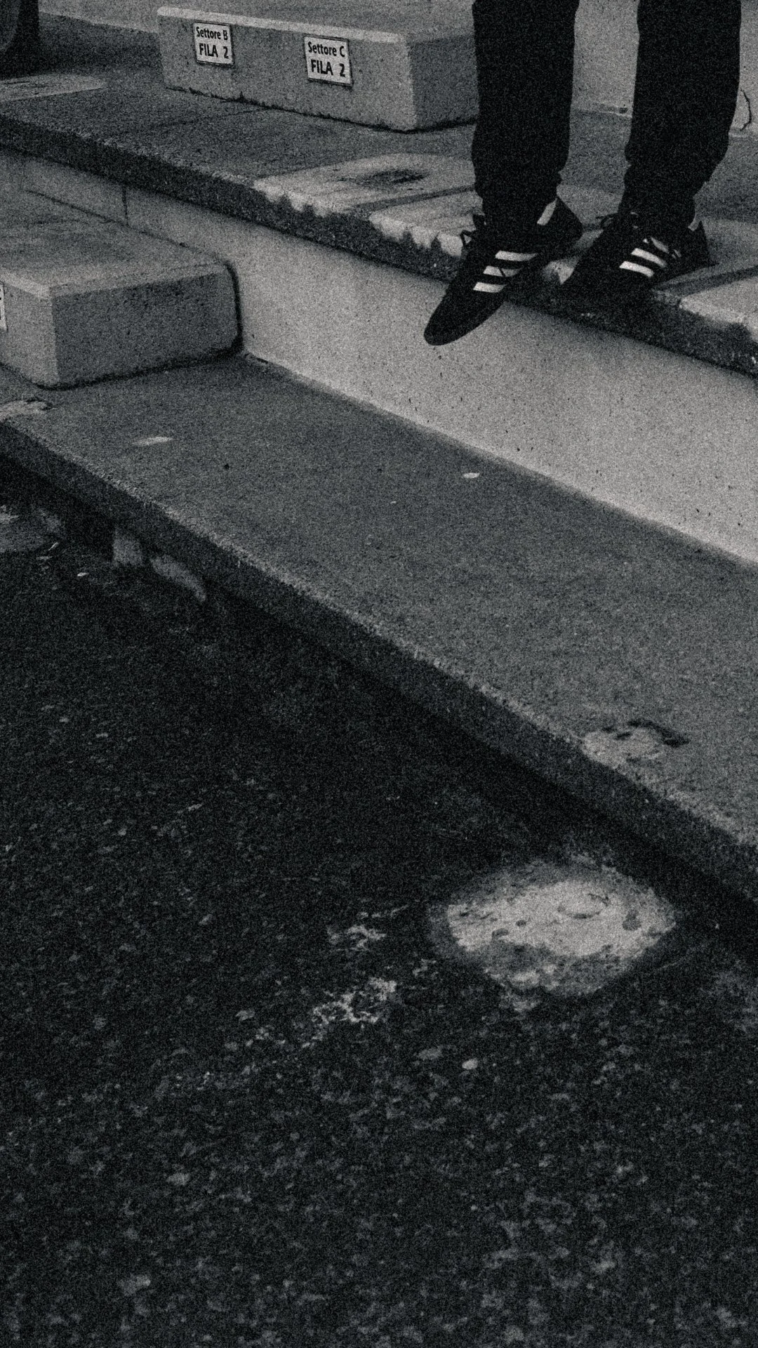 A person's feet wearing black sneakers with white stripes, standing on the edge of a sidewalk near a curb. There are signs indicating parking sections labeled 'Settore B FILA 2' and 'Settore C FILA 2' on a wall in the background.