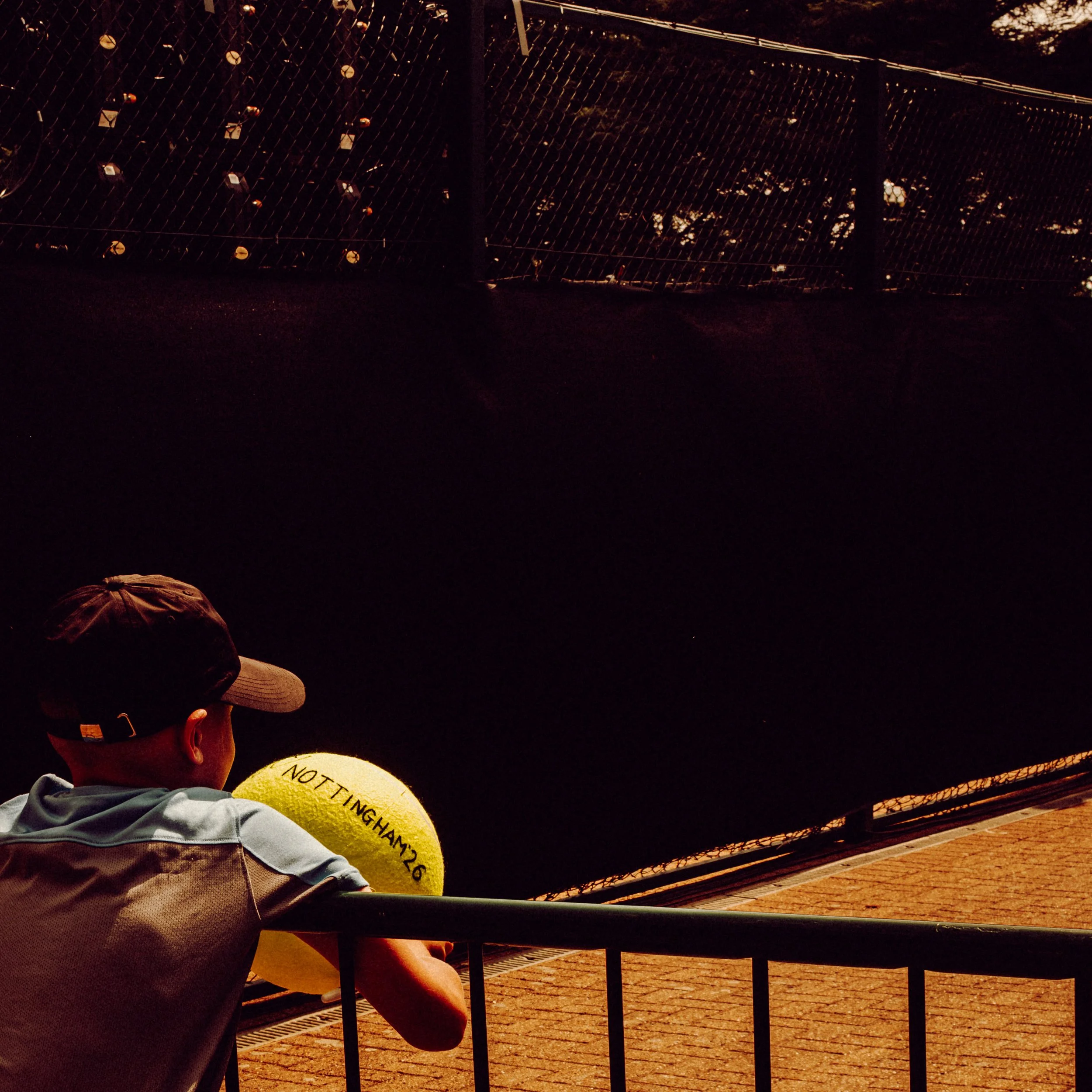 A person wearing a wide-brimmed hat and a jacket, sitting at a table outdoors at night. The person is holding a tennis ball with writing on it. There is a chain-link fence in the background with small lights strung along it.