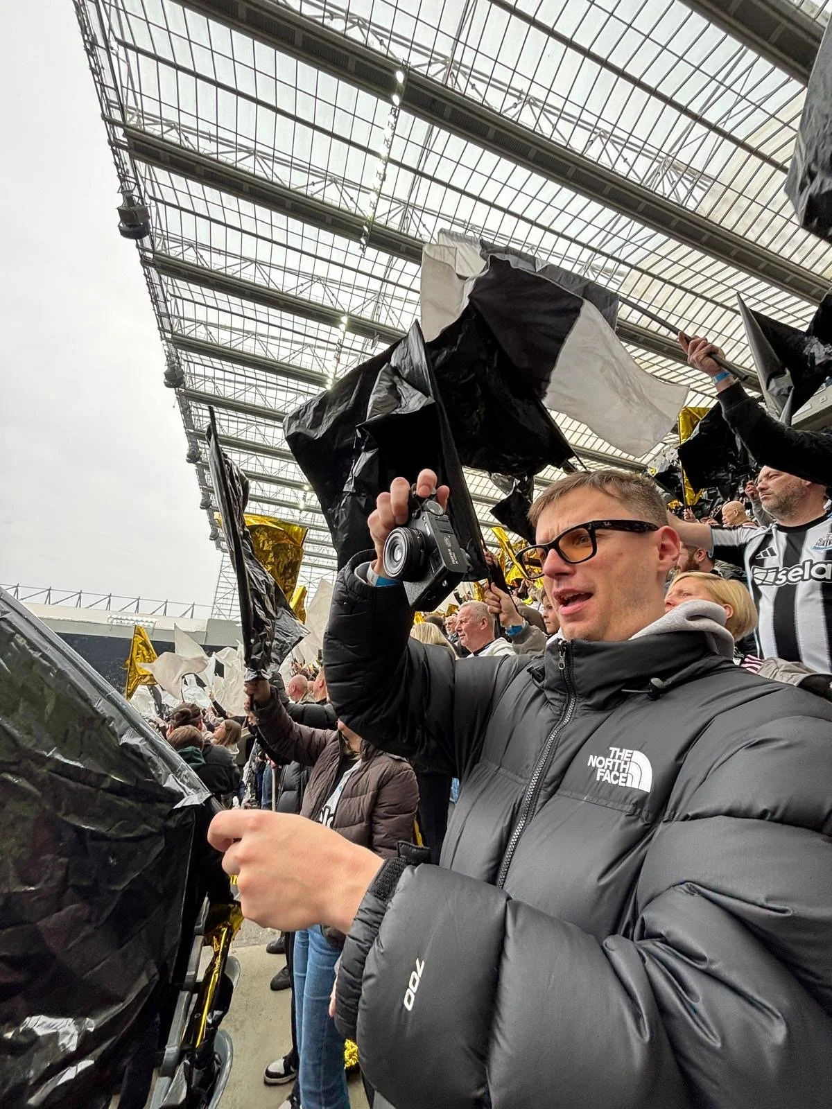 Crowd of sports fans at a stadium, waving black and gold flags, with some wearing team apparel. Overcast sky, large stadium roof in the background, person in the foreground holding a camera, wearing glasses and a gray North Face jacket.