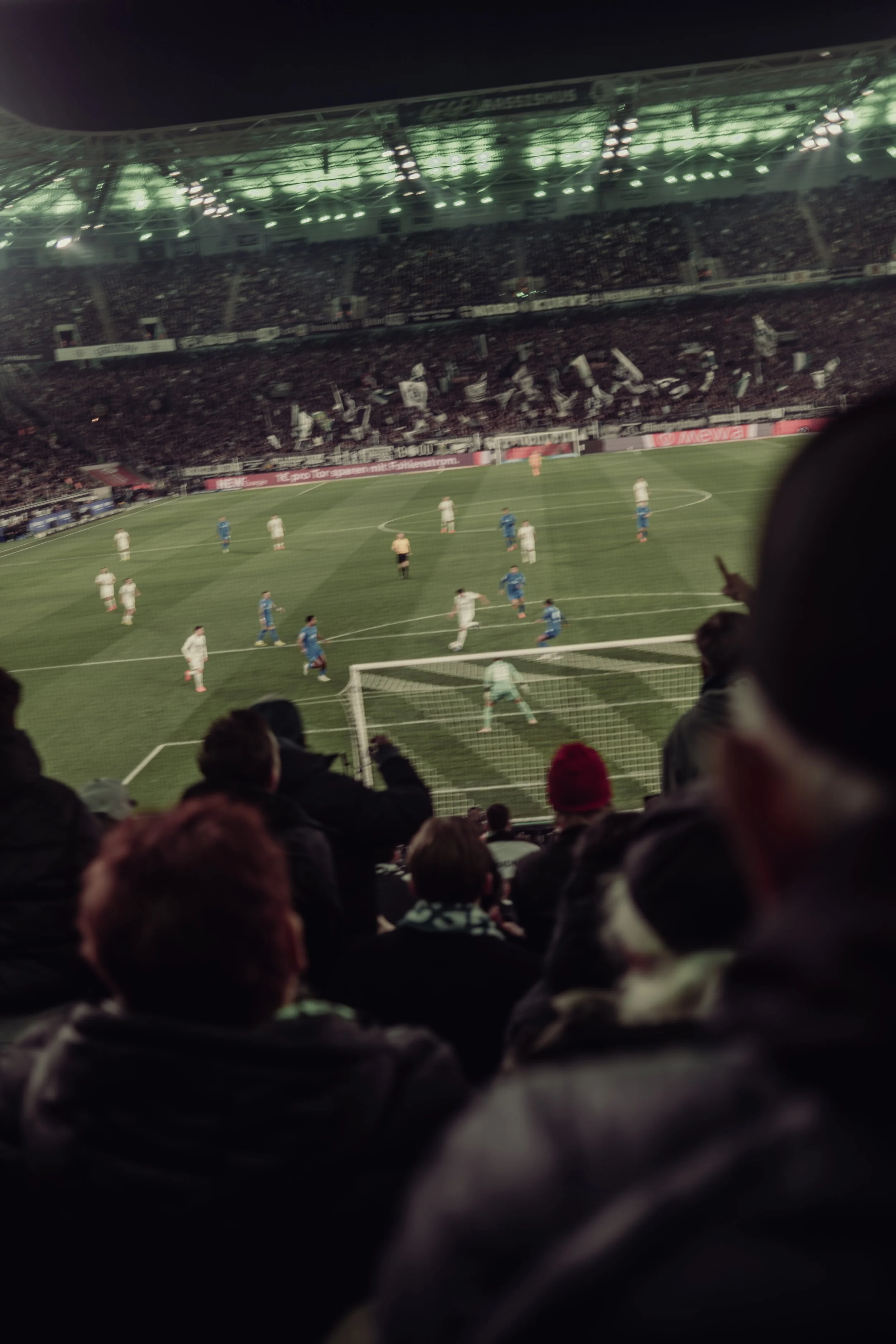 Nighttime scene at a soccer stadium filled with spectators. The field is visible with players in white and blue uniforms, and the stadium is illuminated with bright lights, with fans waving flags and cheering.