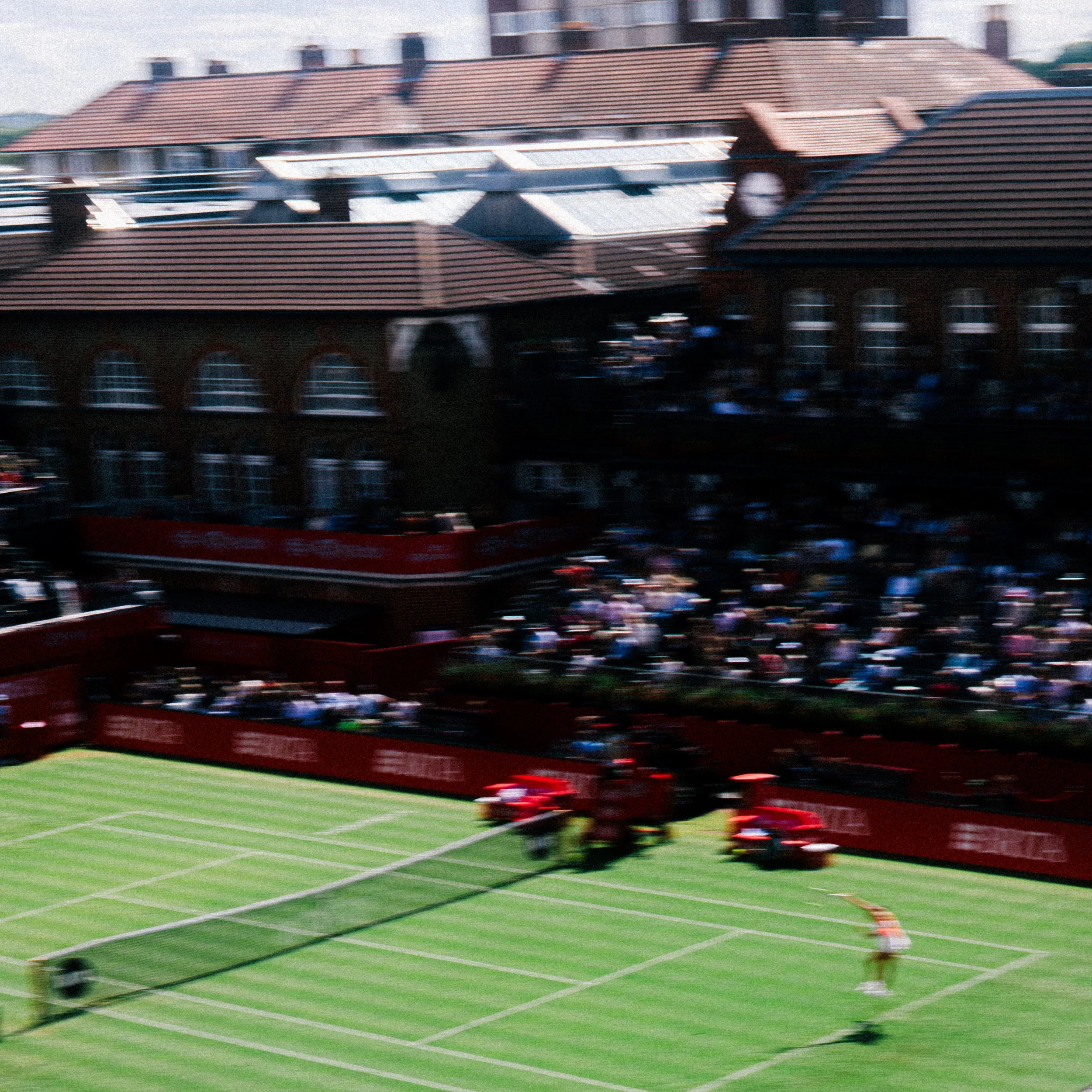 A blurred photo of a sports stadium or field, possibly with a running track, with some buildings or houses visible in the background.