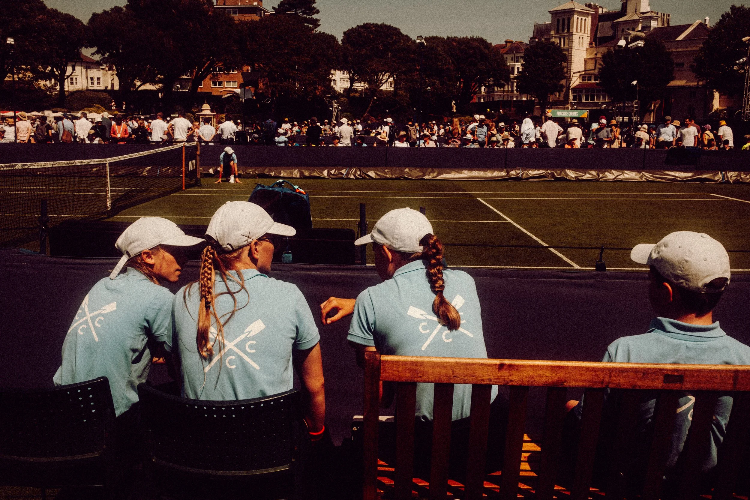 People sitting in front of a tennis court watching a match, with many spectators gathered along the fence and some players and officials on the court, trees, and buildings in the background.