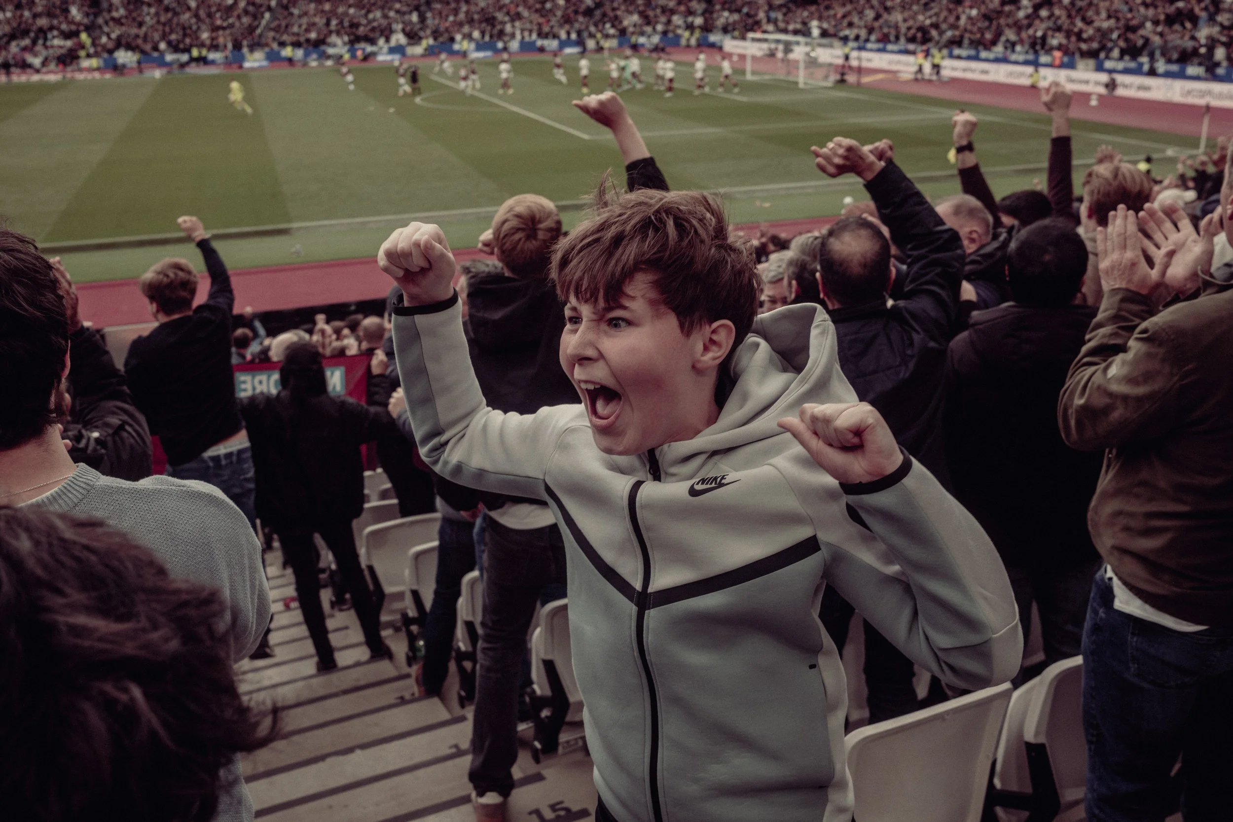 Excited young boy in a white Nike hoodie celebrating at a soccer stadium with spectators cheering in the background.