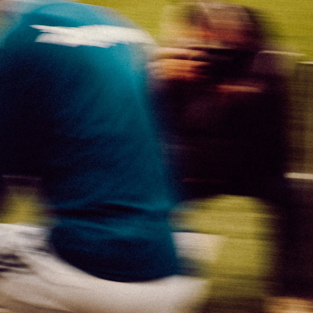 Blurred image of a person in a blue shirt and another person in a black shirt, with a grassy background.