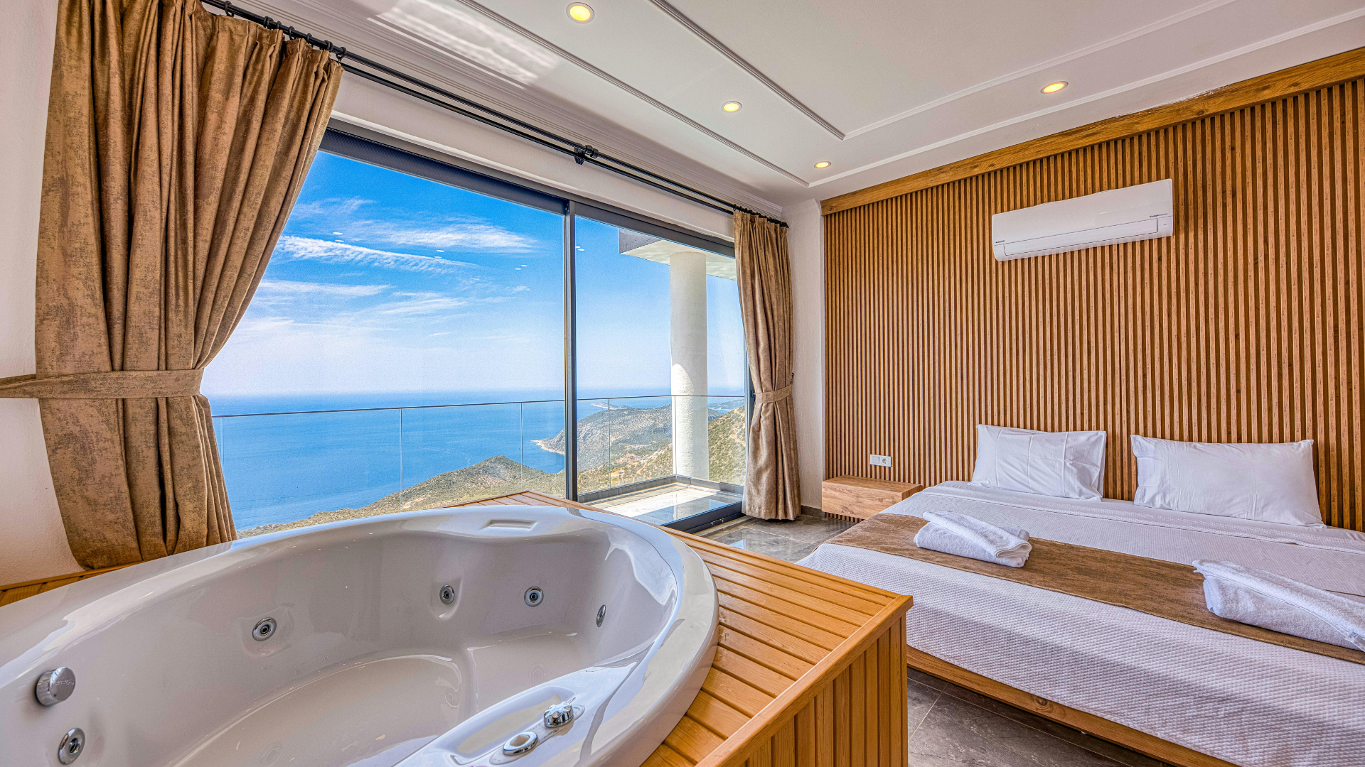 A hotel room with a large window view of the ocean and mountains, a bed with white linens, a folded towel, and a wooden jacuzzi in the foreground.