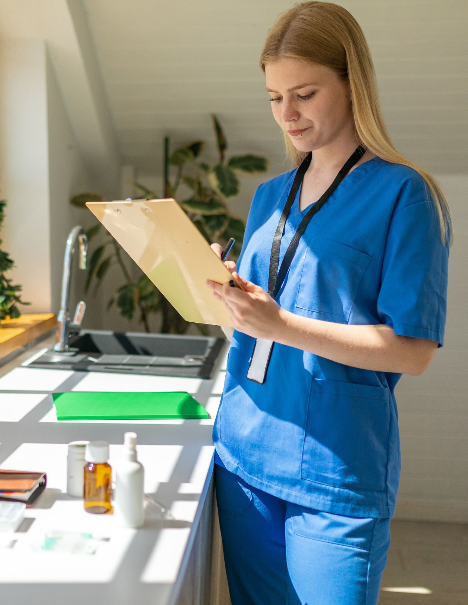 A woman in blue scrubs standing at a white table, holding a clipboard and writing. The table has bottles, a notebook, and various medical supplies. The background shows a plant and a ceiling with wood panels.