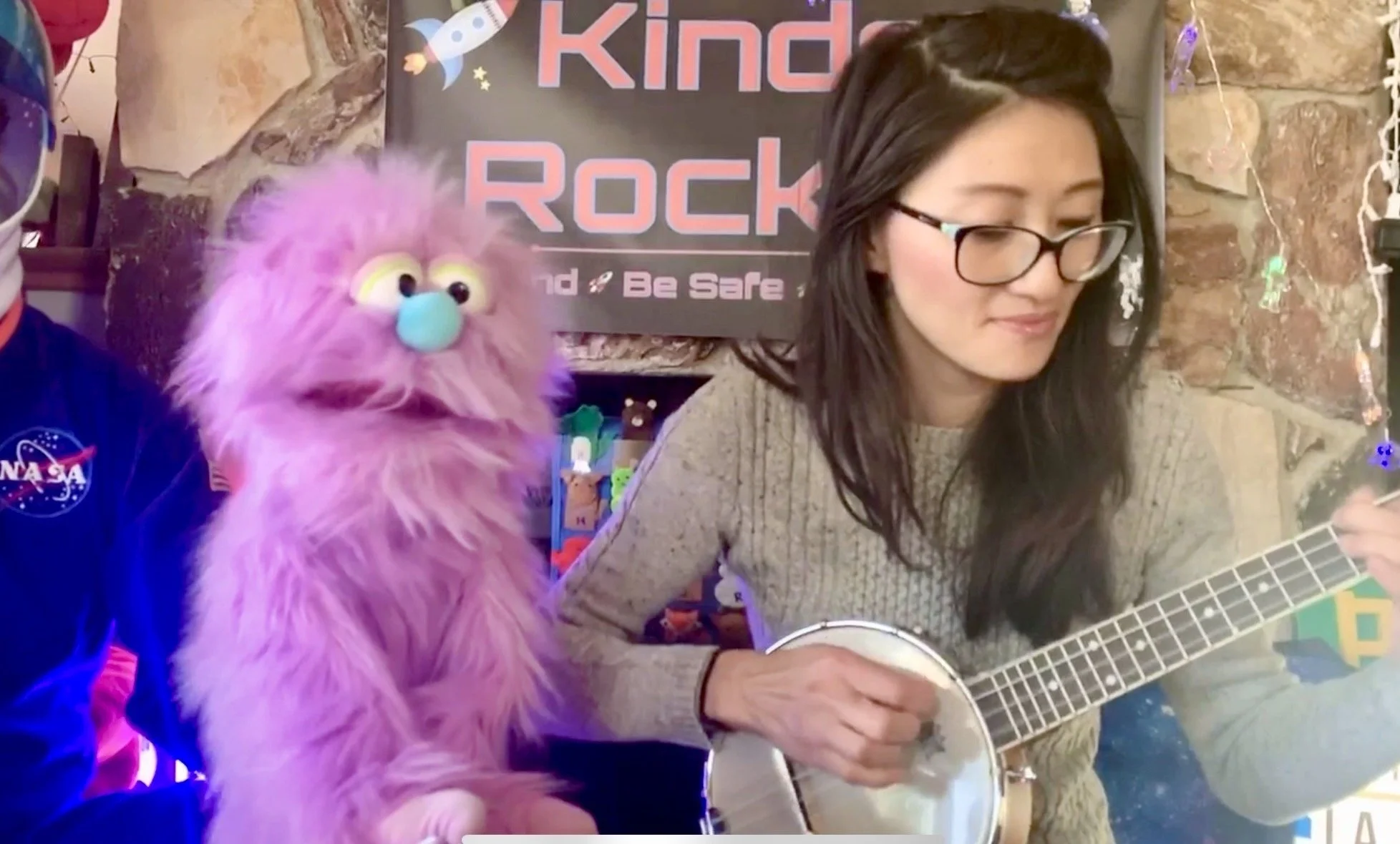 A woman with dark hair and glasses is playing a banjo-ukulele. Next to her is a purple puppet with big yellow eyes, a blue nose, and fluffy fur. The background features a sign that reads "Kinder Rockets" with an illustration of a rocket and stars.