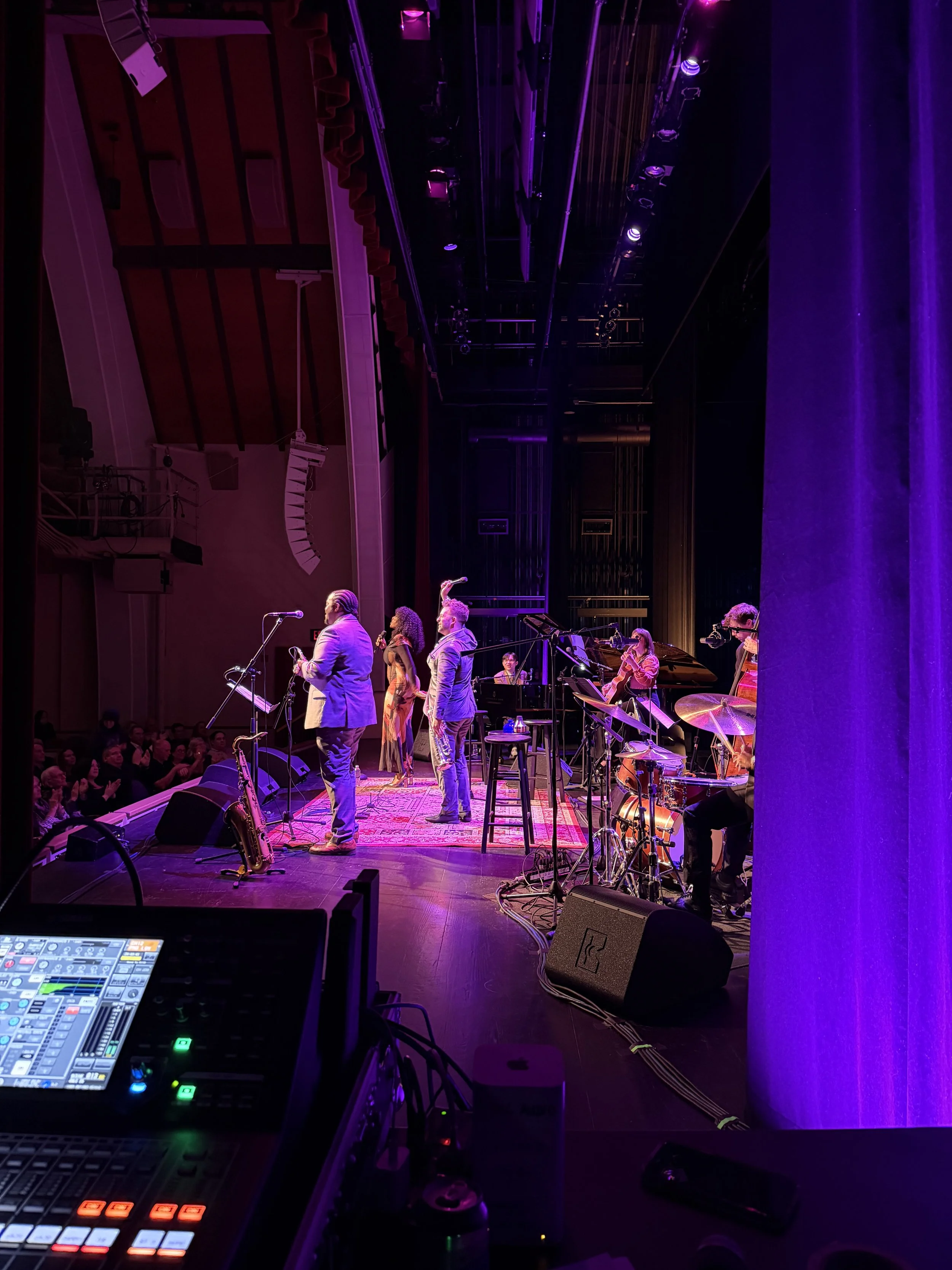 A stage with musical performers and a sound mixing console visible in the foreground, dim purple lighting, and an audience in the background.