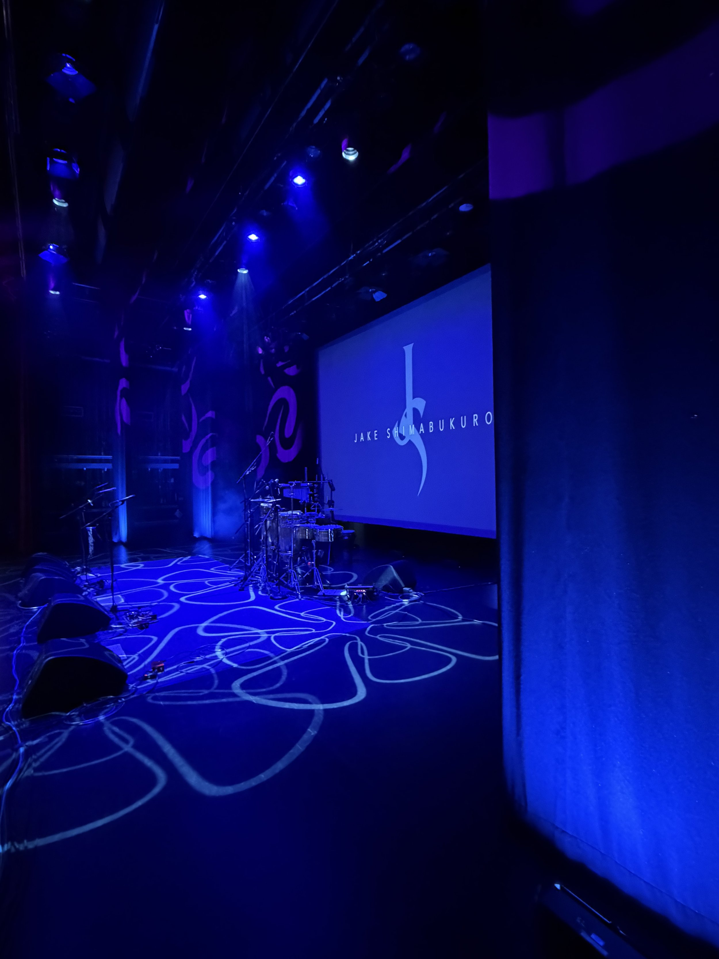 Dimly lit stage with blue lighting, musical instruments, and a large screen displaying the text 'Jake Shimabukuro' with a stylized musical note logo.