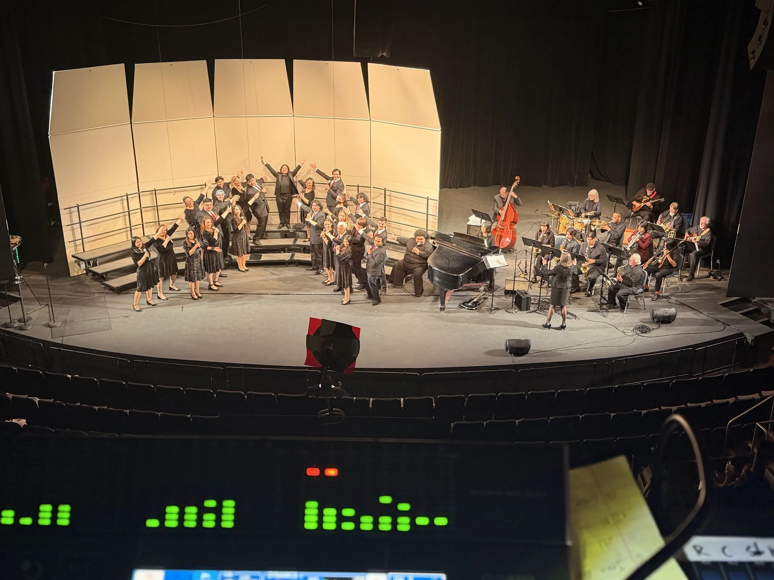 A stage with musicians and dancers performing, and a large beige screen or backdrop behind them. Audience seating is visible in the foreground, and a sound mixing console is in the lower part of the image.