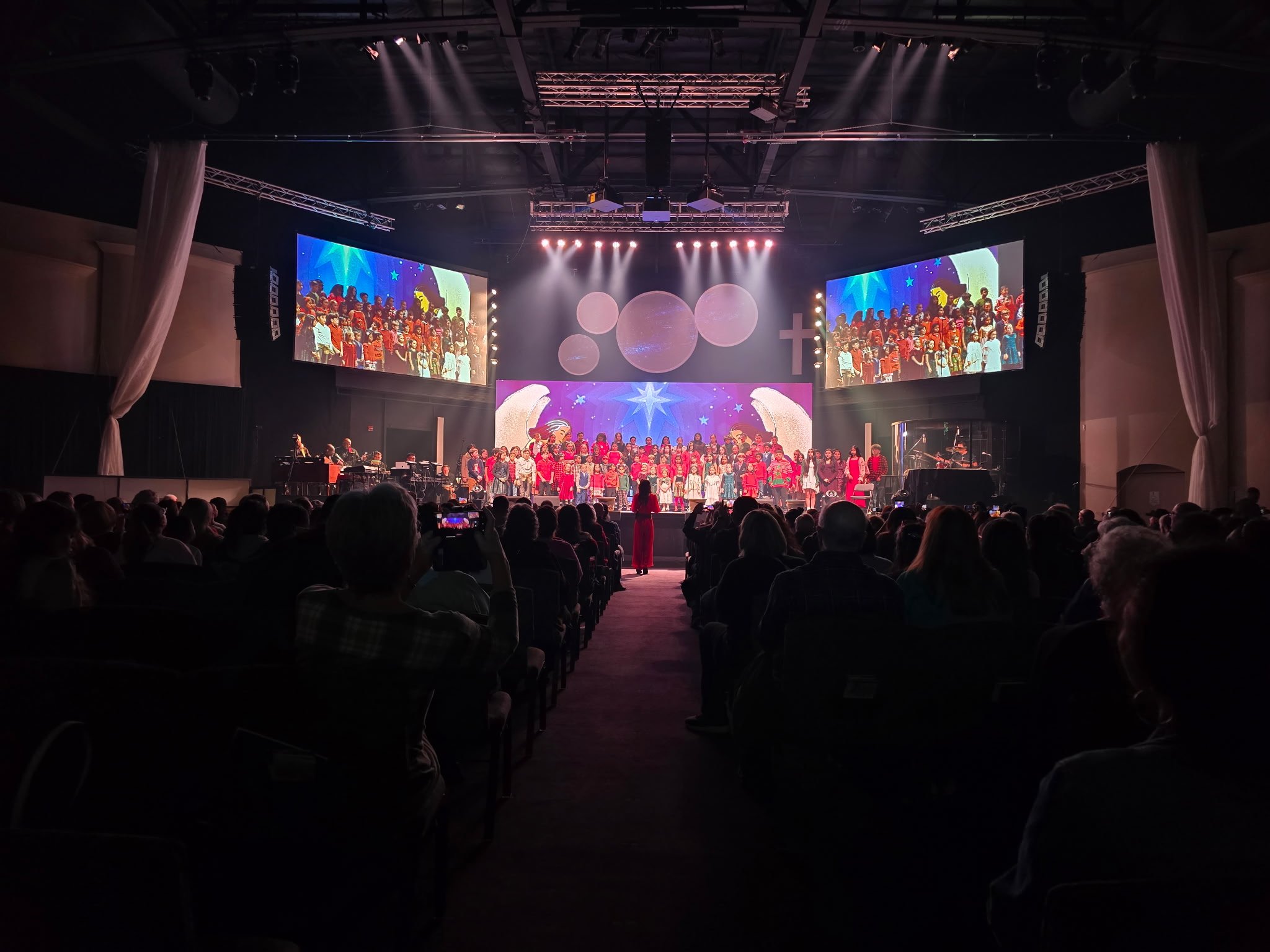 A large group of children performing on stage during a choir or musical concert, viewed from the audience with two large screens displaying the same scene on either side of the stage.