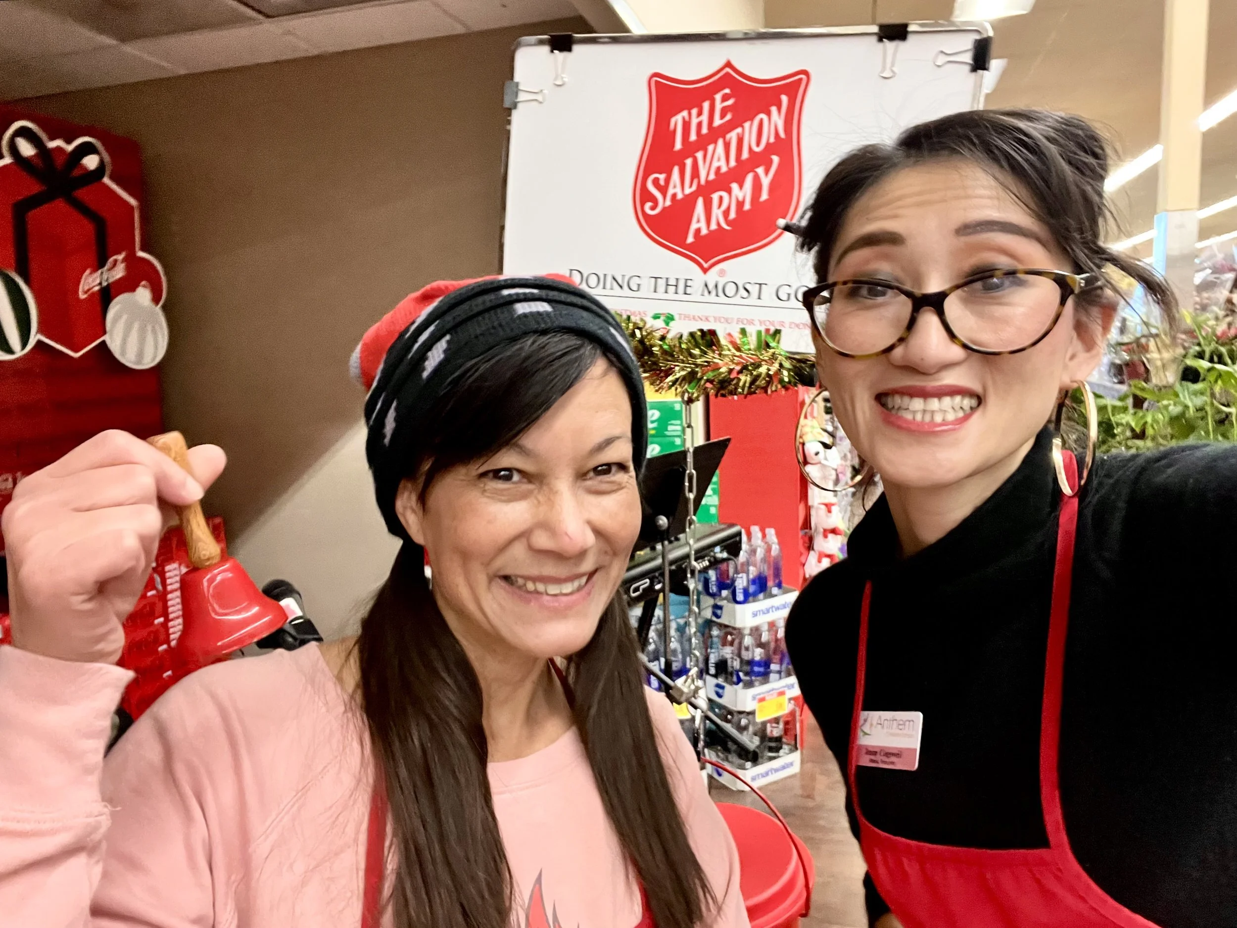Two women smiling inside a store, one wearing glasses and the other wearing a hat and a pink shirt. A Salvation Army sign is visible in the background.
