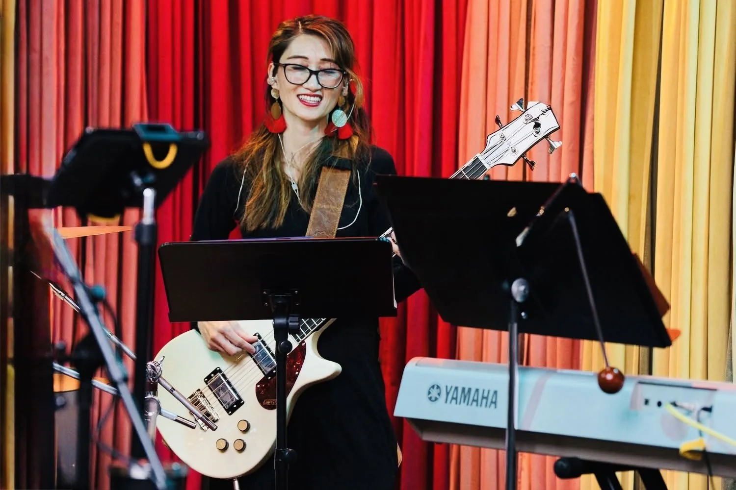 A woman with glasses, earrings, and long hair holds a cream-colored electric guitar on stage with red and yellow curtains in the background. She is smiling and standing in front of music stands and equipment.