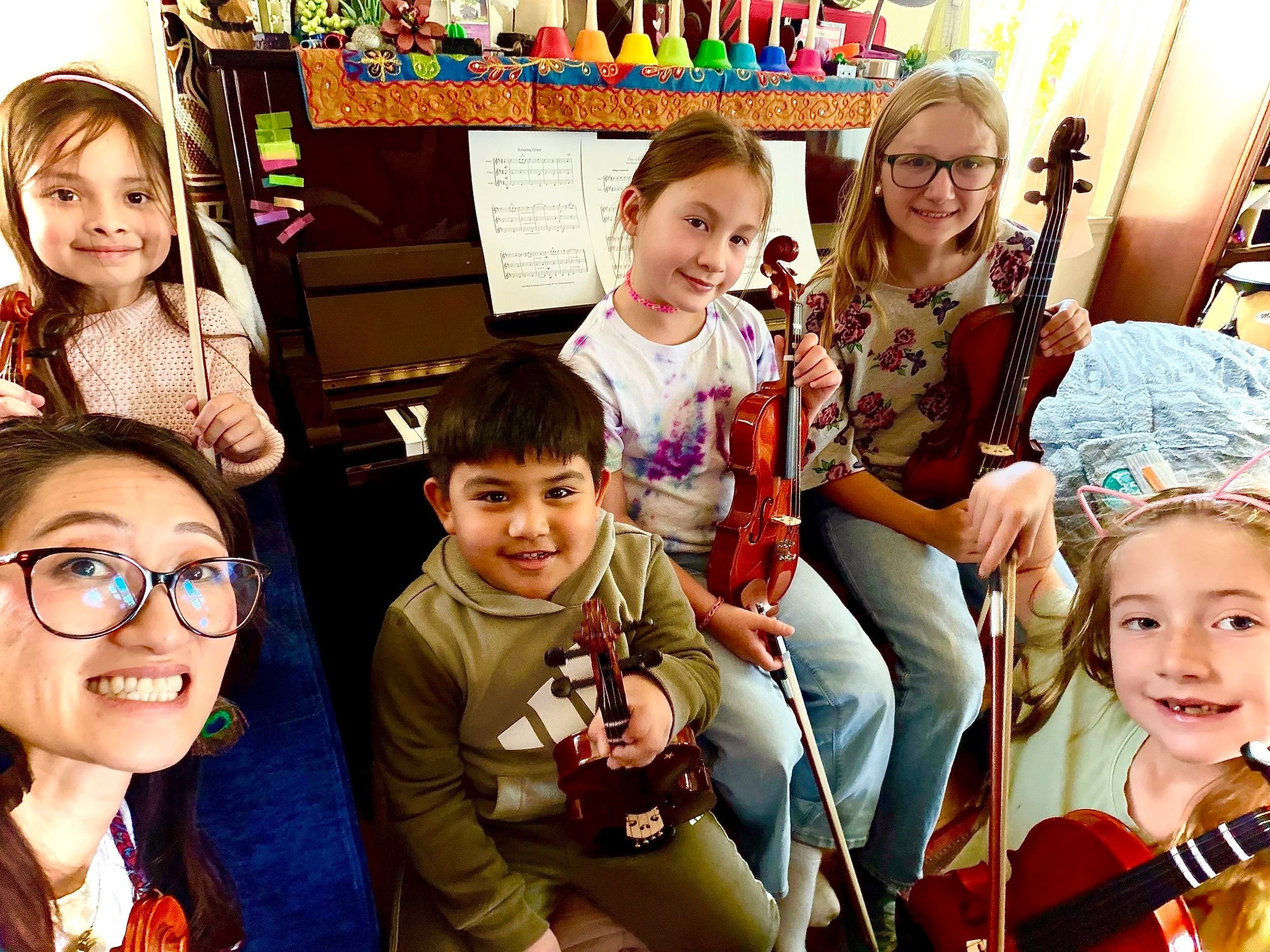 Group of children and an adult woman indoors, holding violins and smiling, with a piano, sheet music, and colorful decorations in the background.