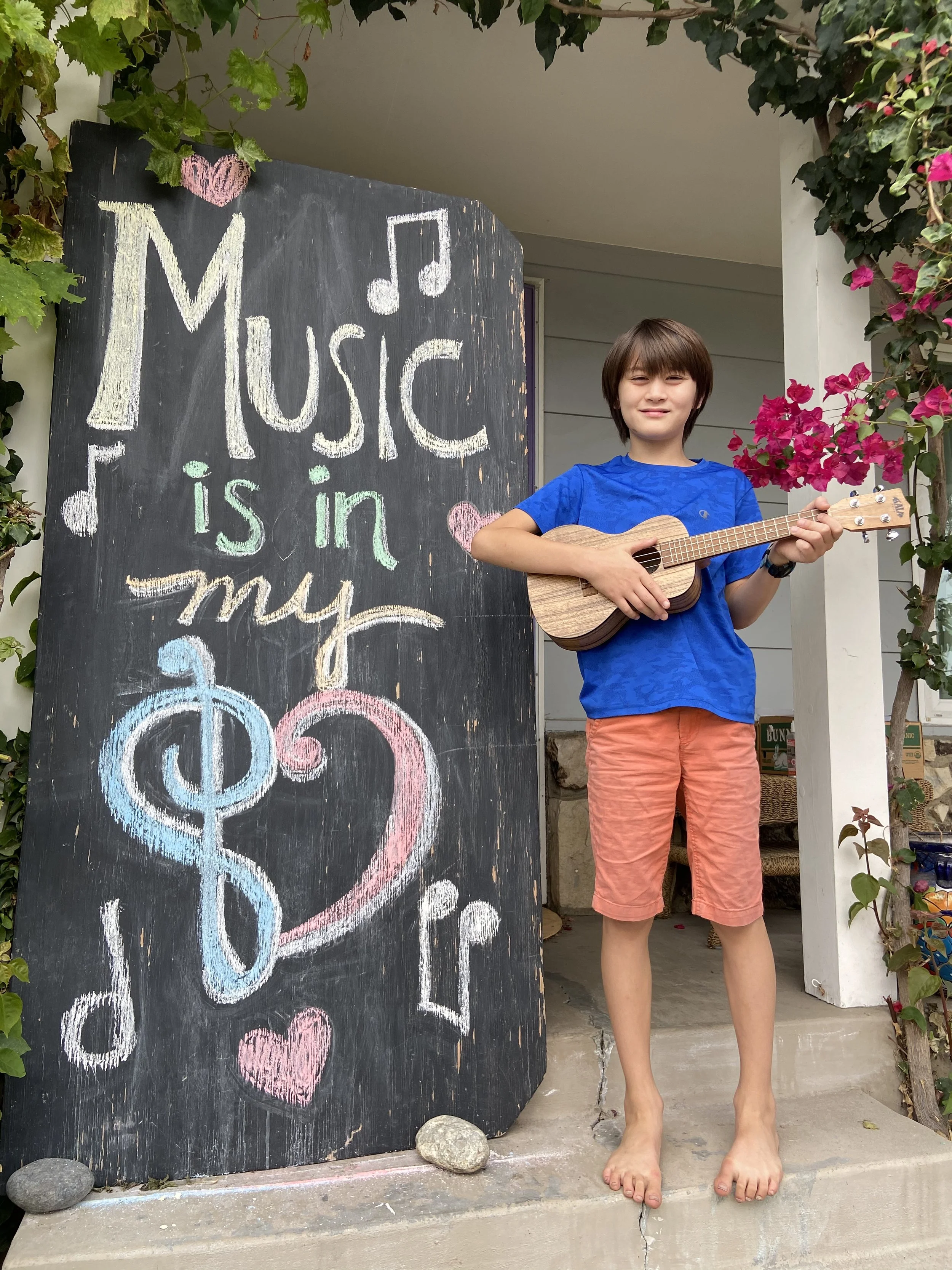 A young boy holding a ukulele standing next to a chalkboard with colorful chalk writing about music, surrounded by greenery and pink flowers.