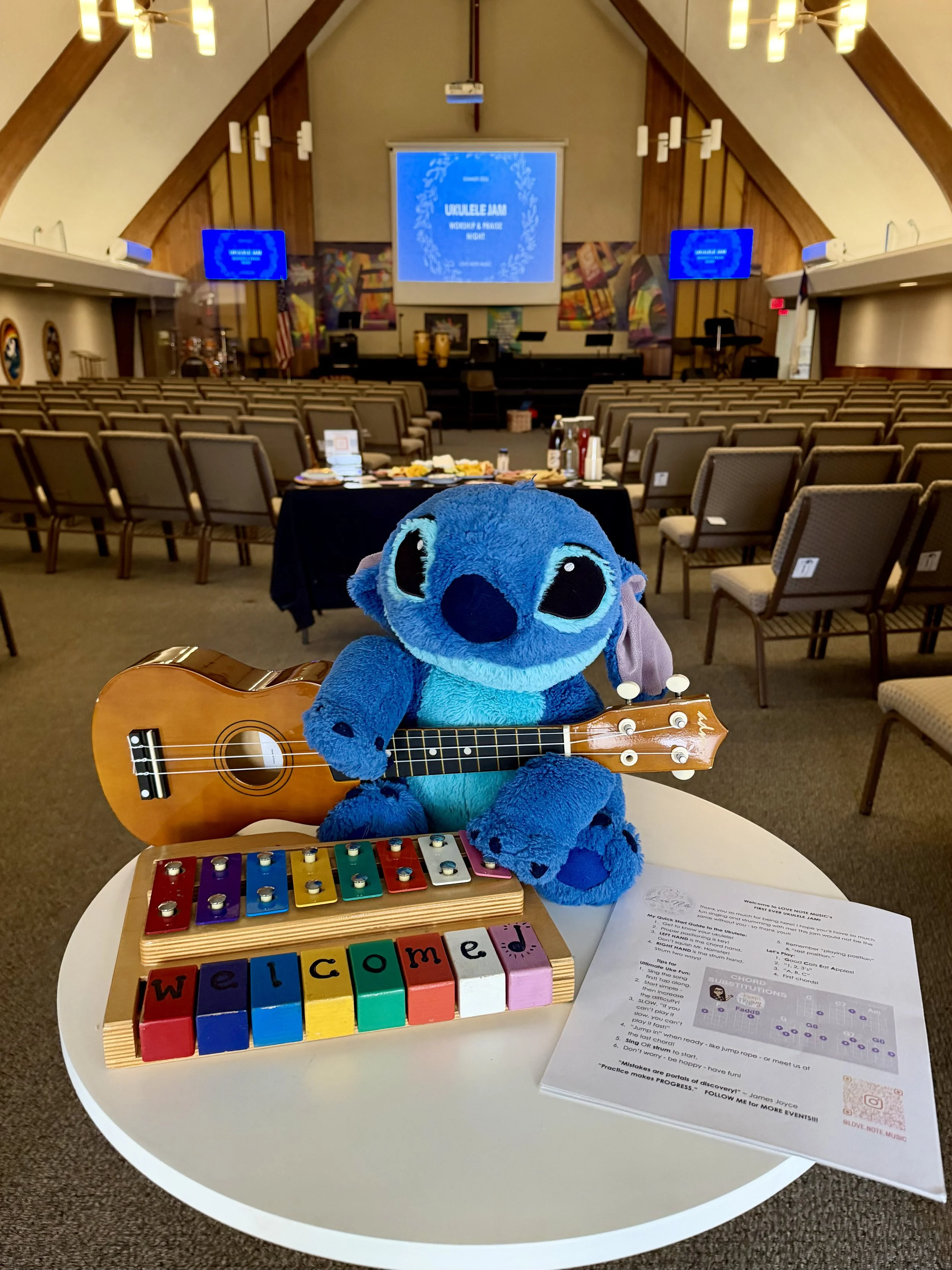 A plush blue Stitch toy holding a small acoustic guitar, sitting on a white table with a colorful wooden 'Welcome' block puzzle, sheets of paper, and small musical instruments. In the background, there is an empty auditorium with rows of chairs and a stage with a large screen displaying 'Ukulele Jam Worship & Praise Night' and two monitors.