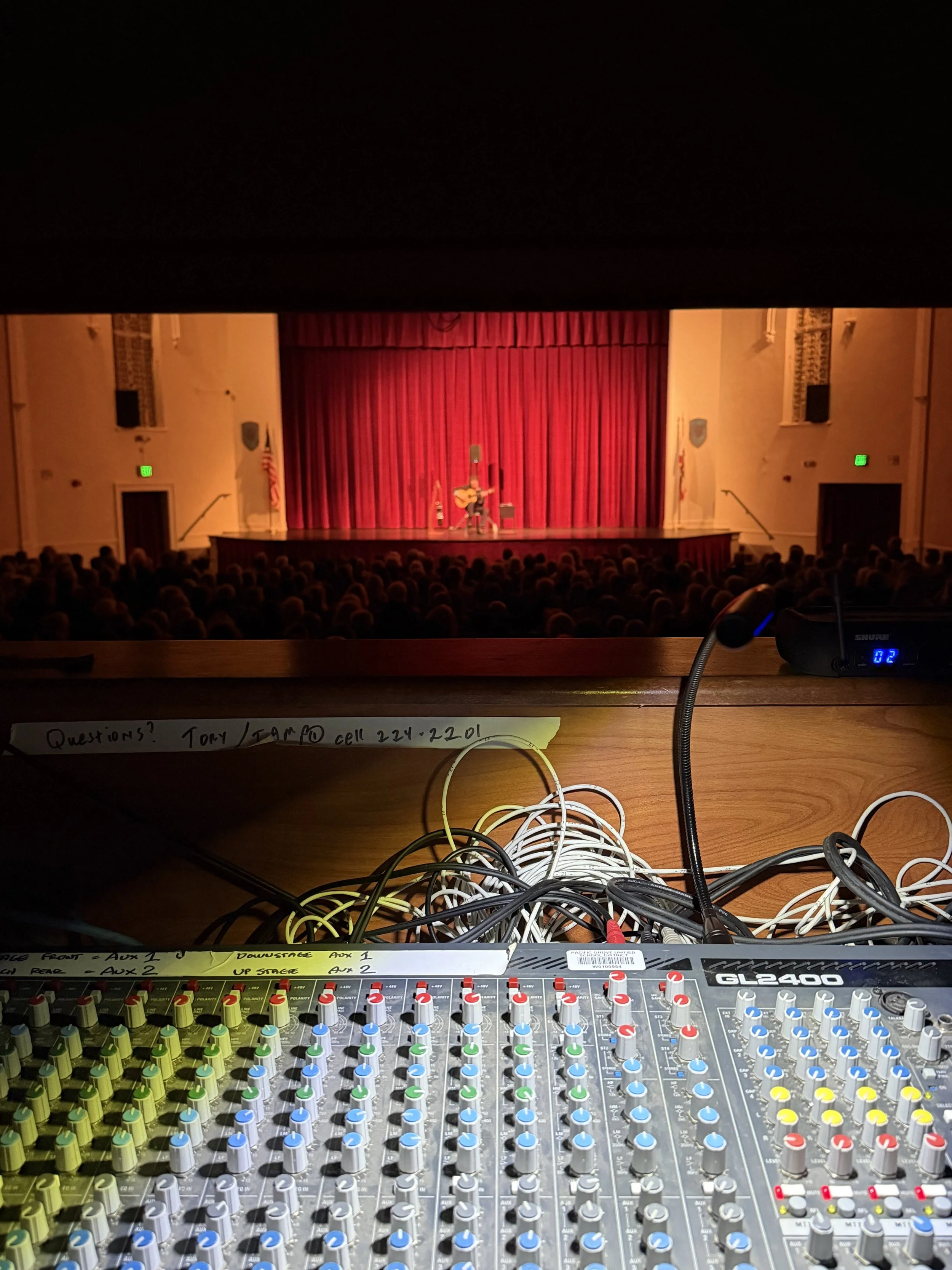 Photo taken from the control booth of a theater or concert hall, showing the audience and stage with red curtains, and a musician playing guitar on stage. In the foreground, there is audio mixing equipment with numerous cables and controls.