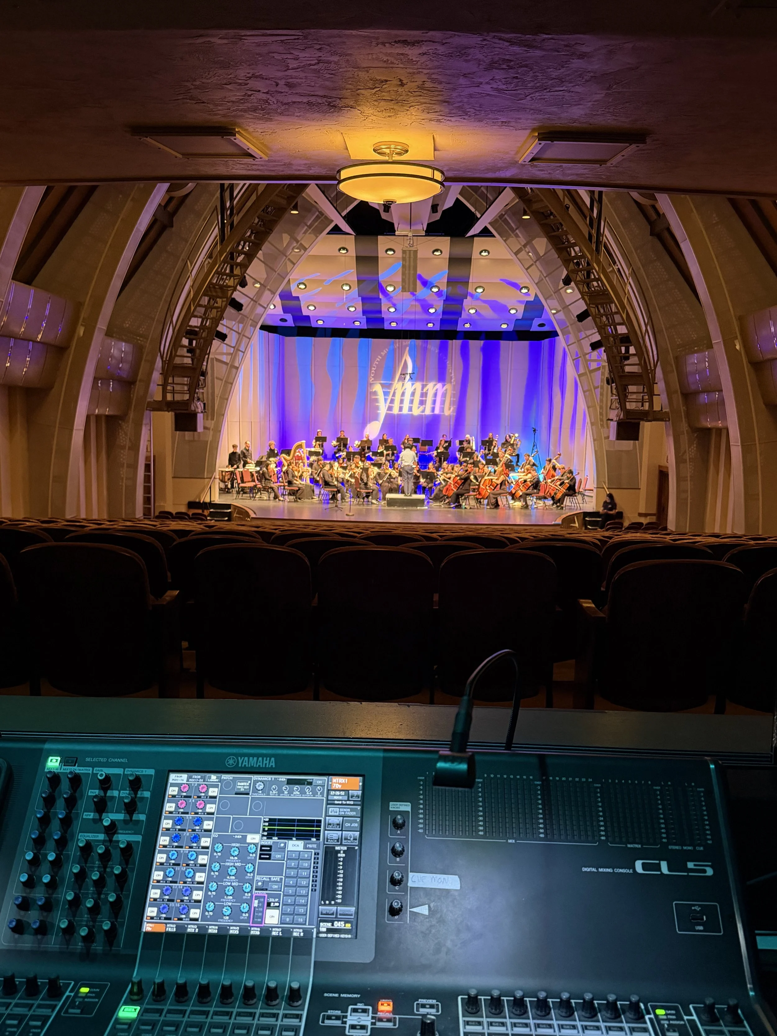 Concert hall stage with an orchestra performing, viewed from behind a digital mixing console in the foreground.