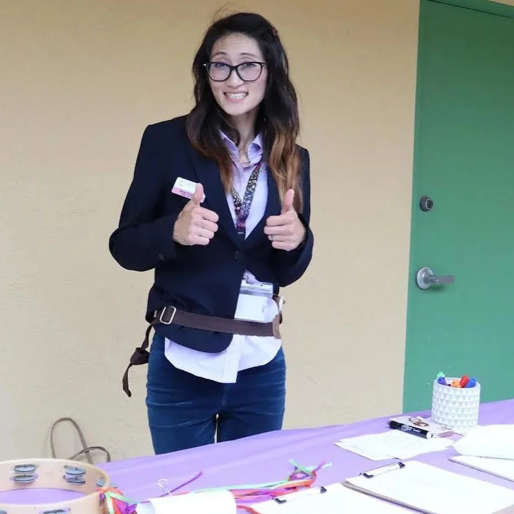 A woman with glasses and long dark hair with light brown tips is smiling and giving two thumbs up. She is wearing a navy blazer, a white shirt, and a lanyard. She is standing behind a table with colorful ribbons, papers, and a small container of markers. The background features a yellow wall and a green door.