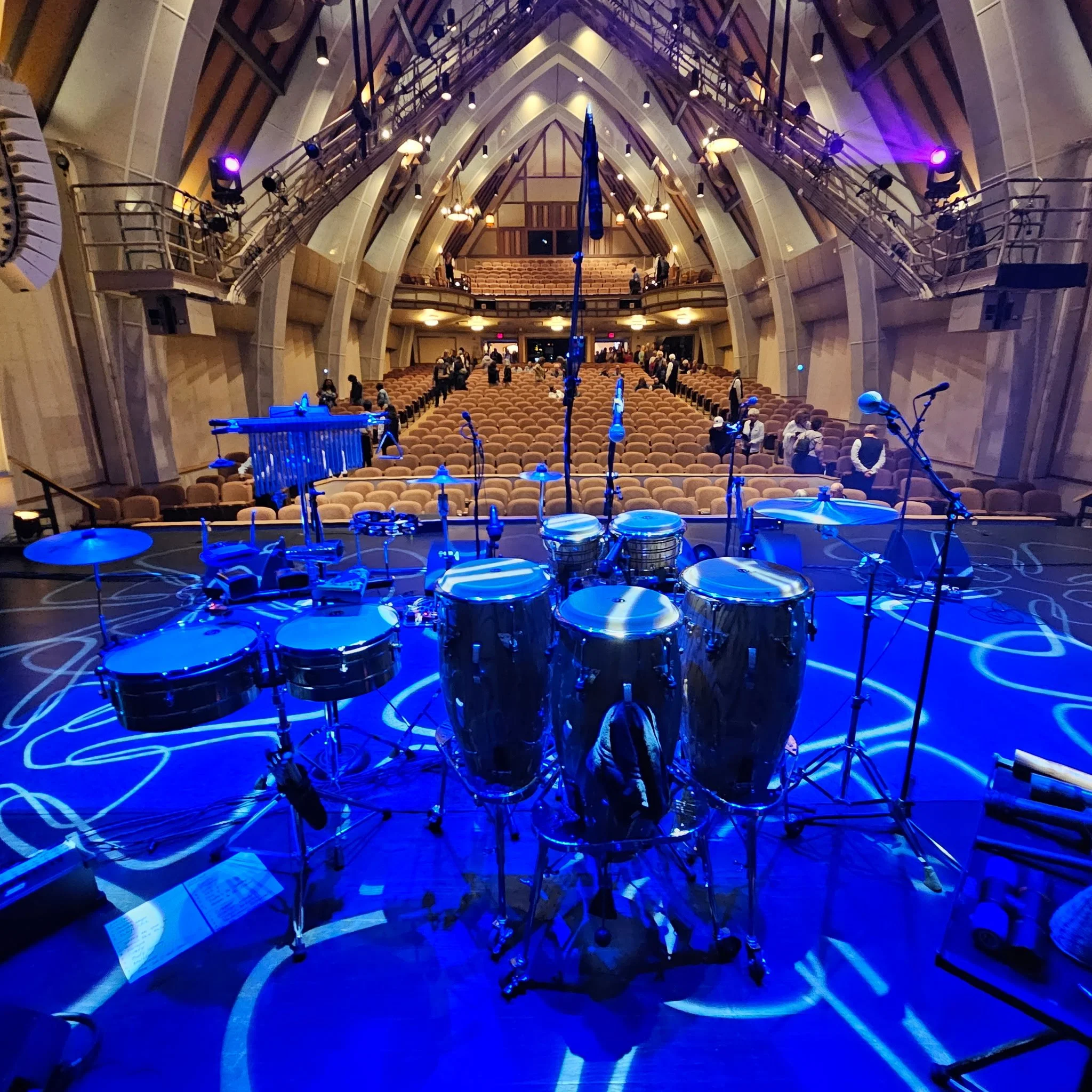 The image shows the interior of a concert hall with rows of chairs, a stage with drums, microphones, and musical instruments, and an arched ceiling with lighting fixtures and stage lights.