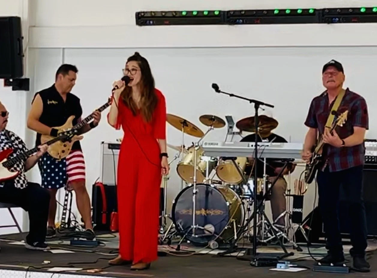 A band performing live on stage with two guitarists, drummer, keyboardist, bassist, and a female singer in a red dress.