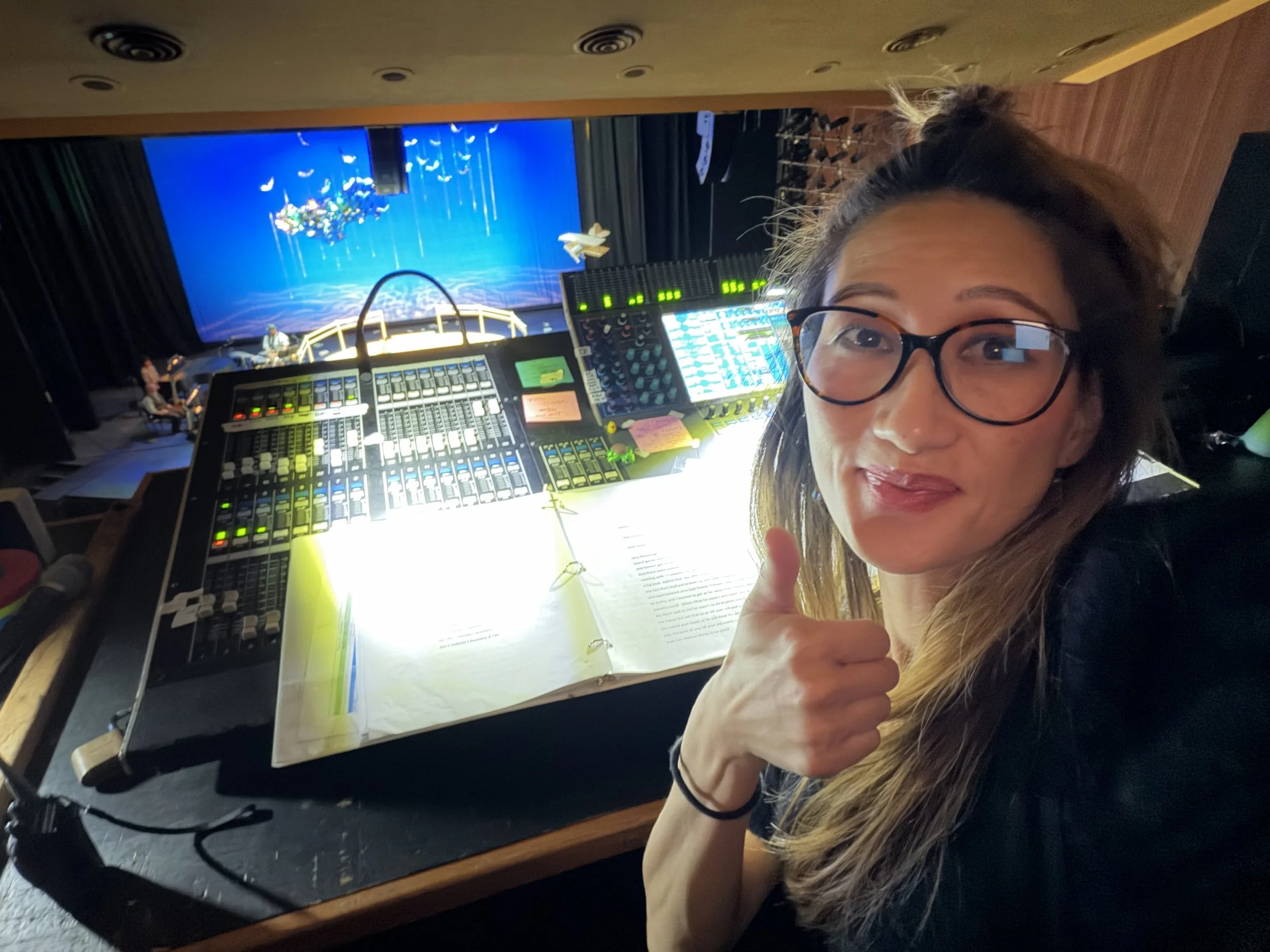 A woman wearing glasses holds a thumb up and smiles while sitting in front of a large music mixer and sheet music, backstage at a theater with a stage in the background.