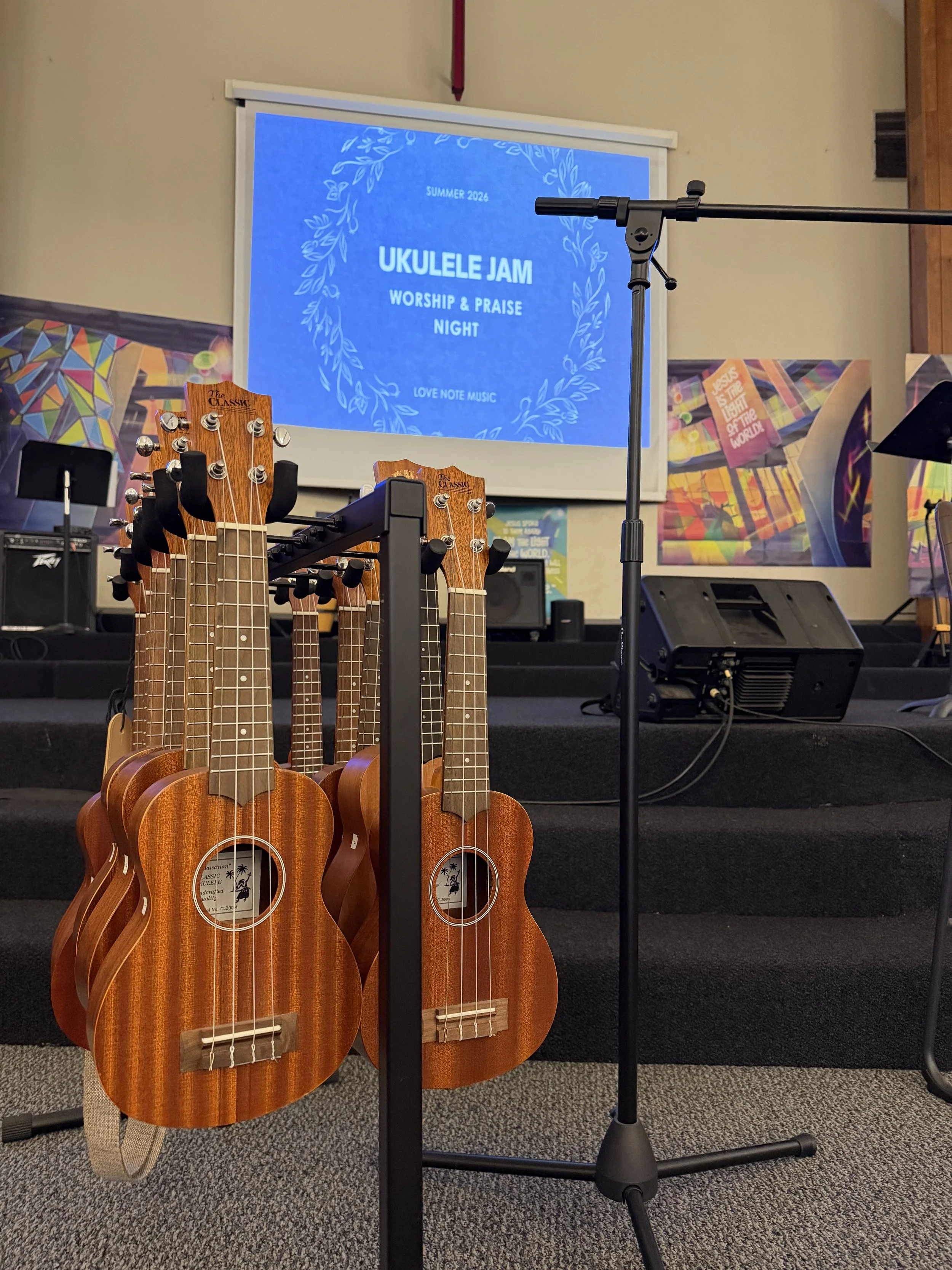 A stage set up for a worship and praise night event called 'Ukulele Jam' with a projected blue screen, four ukuleles on a stand, microphones, and colorful posters in the background.