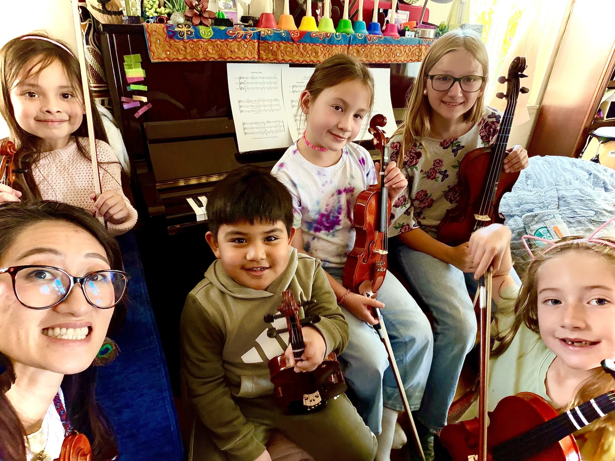 Group of children and an adult smiling, holding violins, in a room with a black piano, sheet music, and colorful decorations in the background.