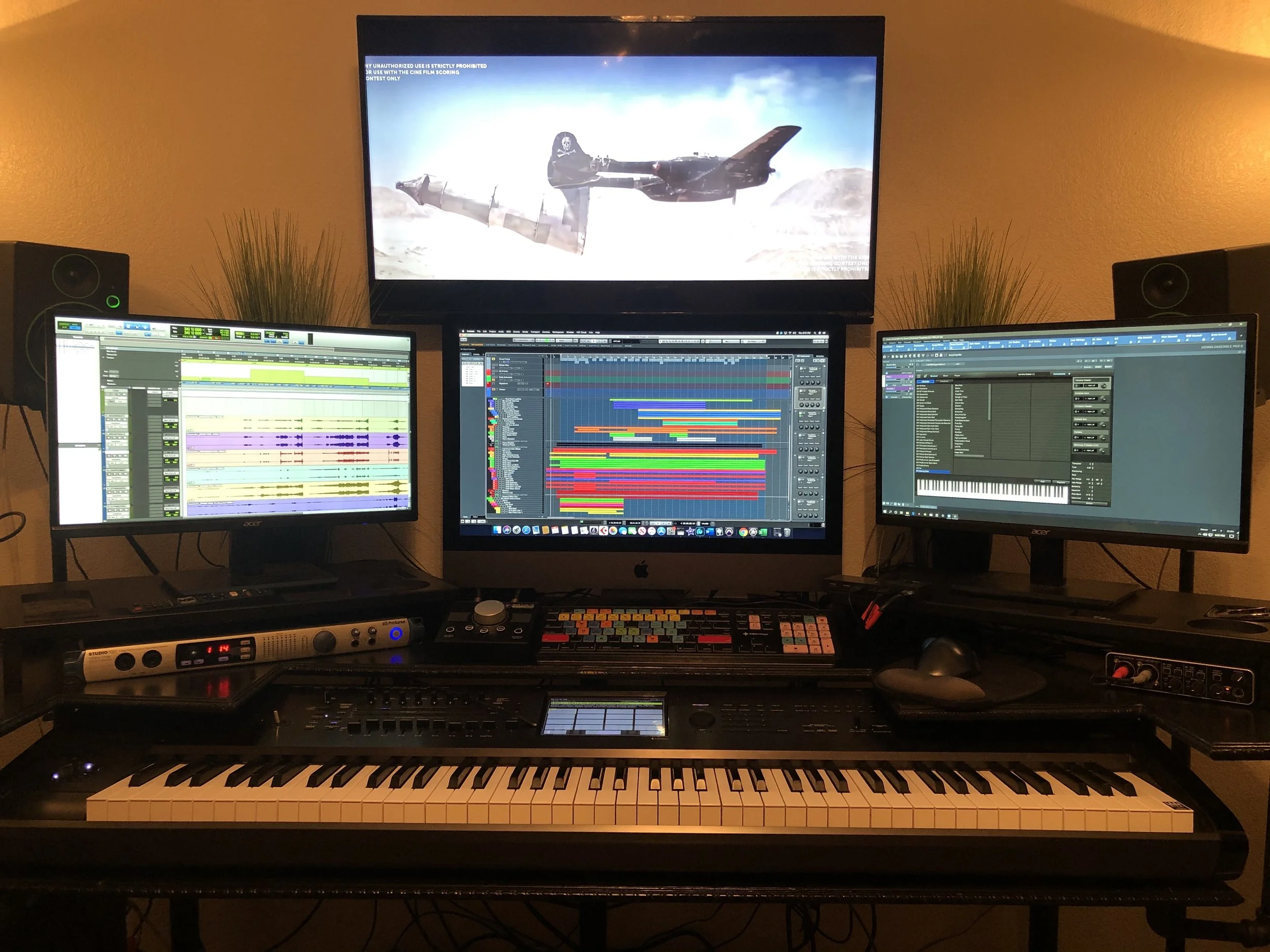 A music production workspace with multiple monitors displaying digital audio workstation software, a keyboard, and audio equipment, with a large screen showing a video game scene featuring a fighter jet in a desert landscape.