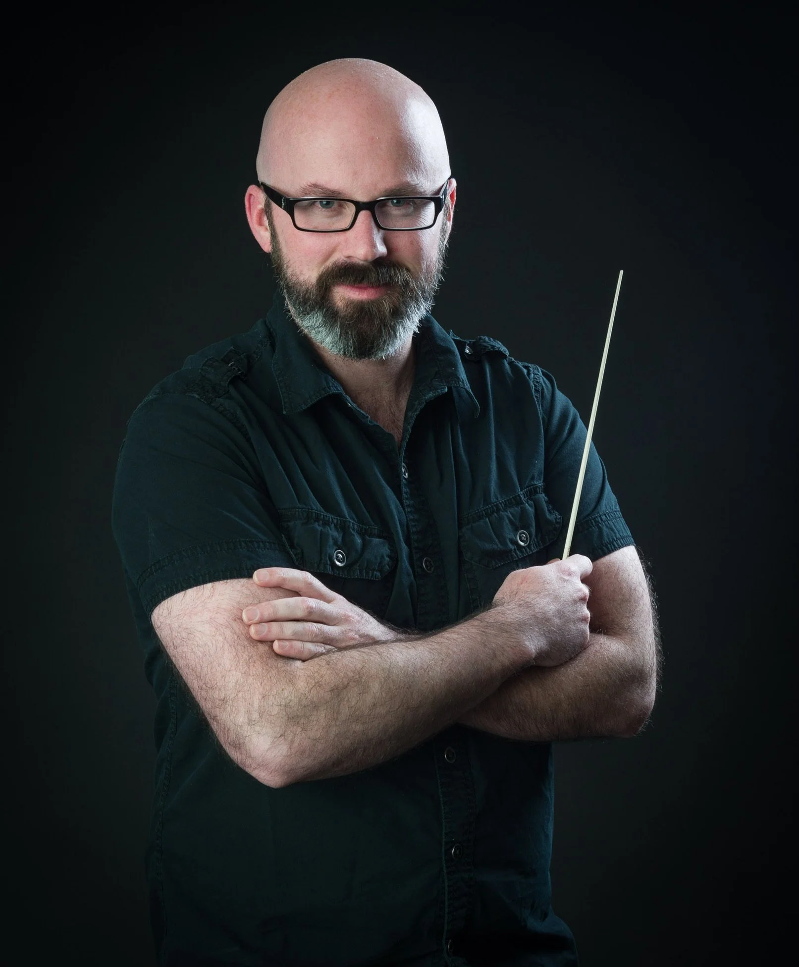 A bald man with glasses and a beard, wearing a black shirt, holding a conductor's baton, standing against a dark background.