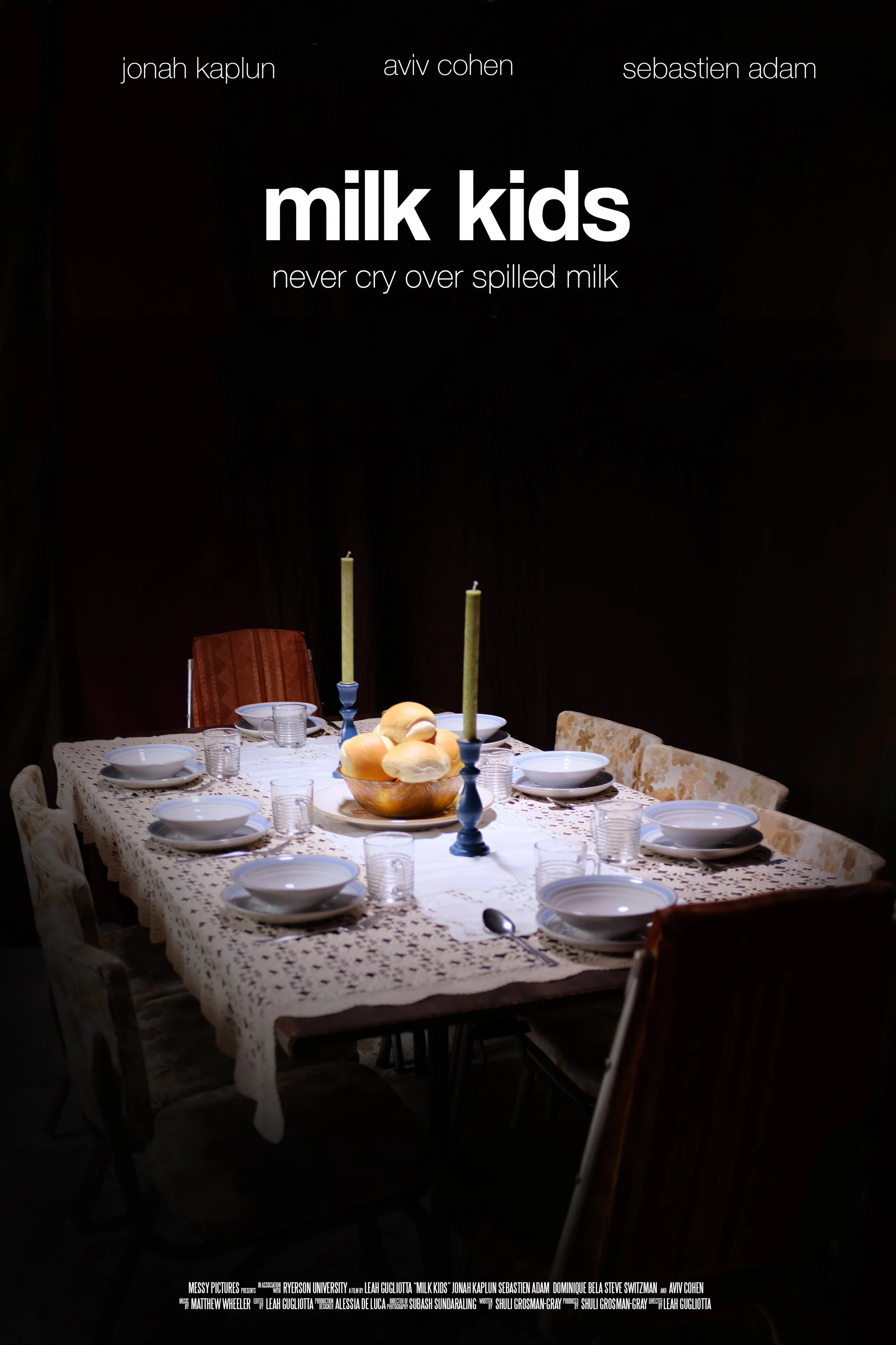 A dining table set for a meal with white plates, glasses, and silverware, covered with a patterned tablecloth. A bowl of bread rolls sits in the center, flanked by two tall candles in candlesticks. The background is dark with a black wall, and there are chairs around the table.