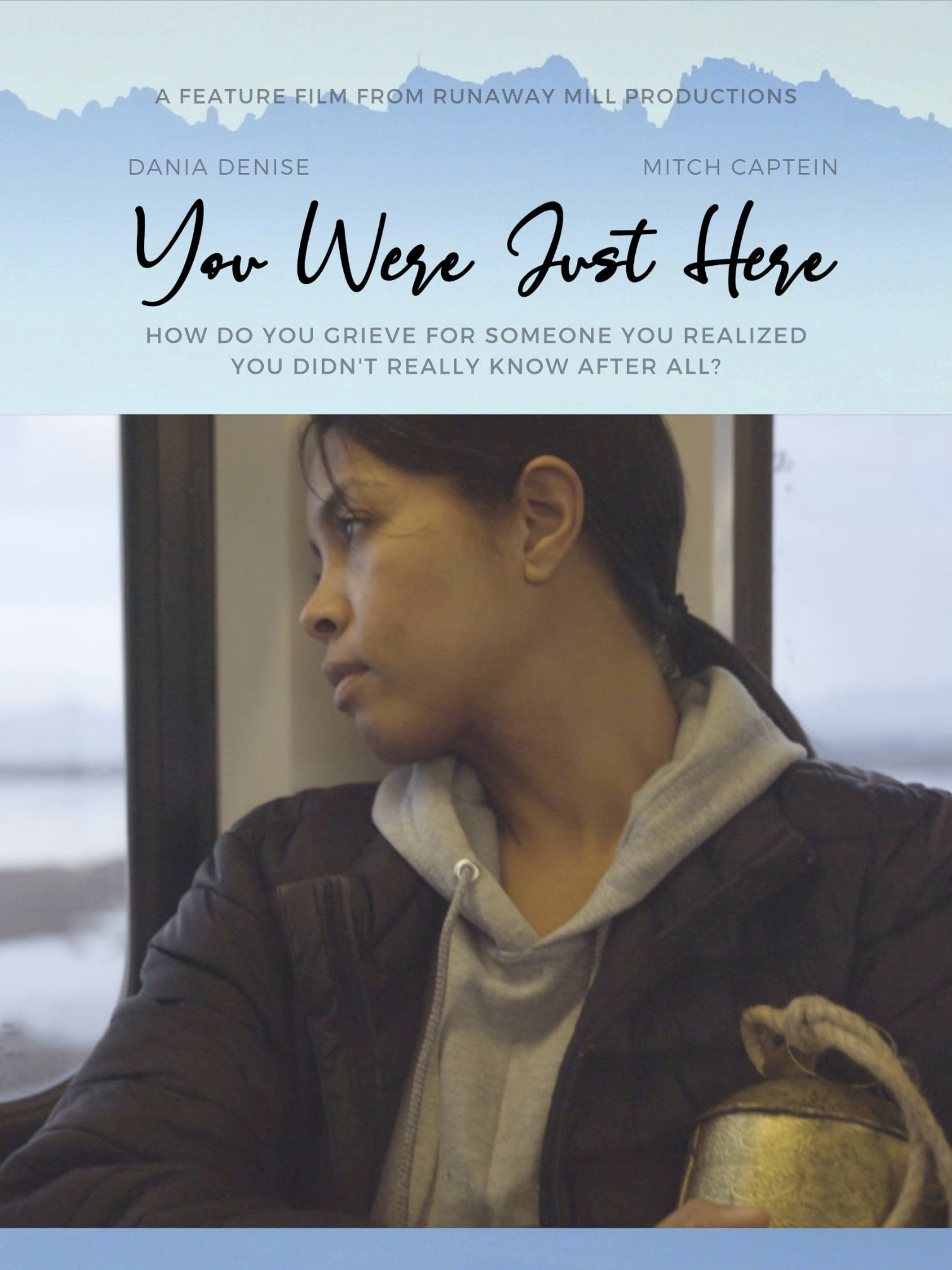 A woman with dark hair wearing a gray hoodie and black jacket, looking out a window, with a view of water and cloudy sky in the background.