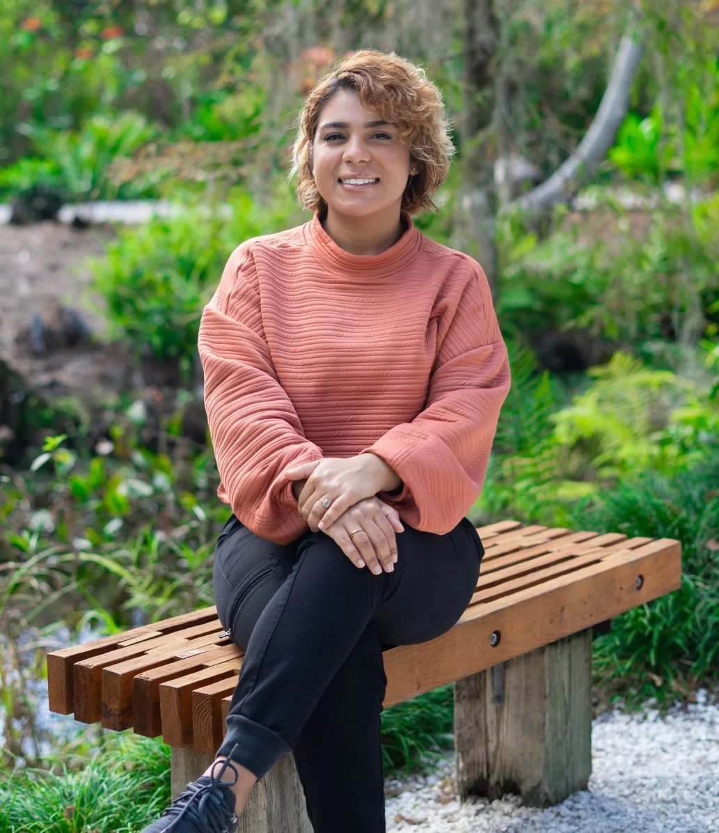A woman with short curly hair, wearing a salmon-colored textured sweater and black pants, sitting on a wooden bench outdoors surrounded by greenery.