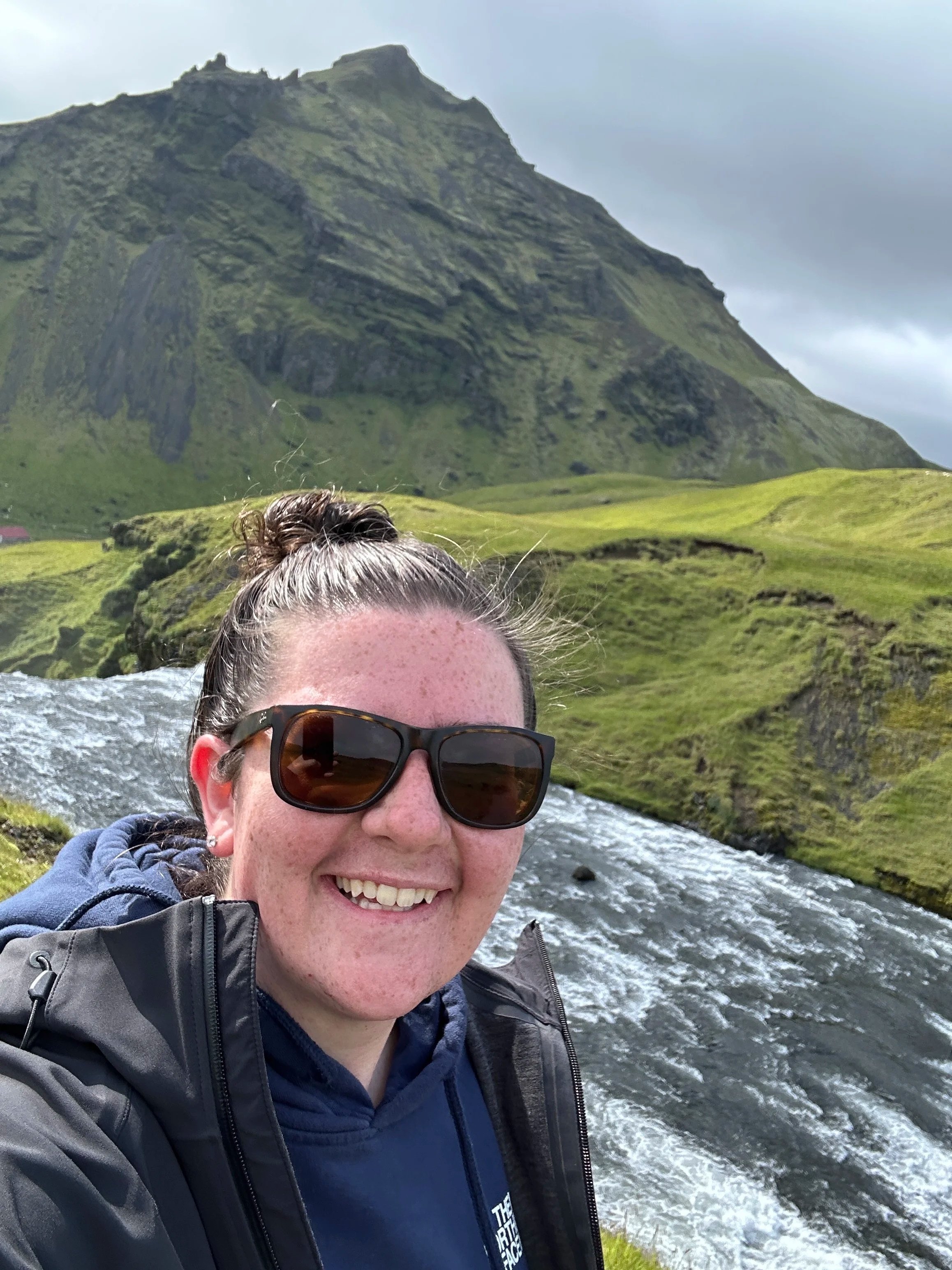 A woman with sunglasses smiling at the camera in front of a flowing river with green hills and a mountain in the background.