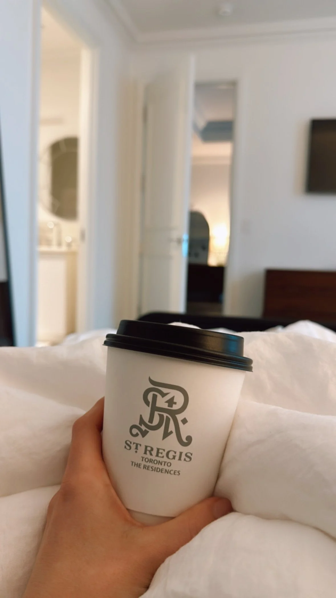 Morning coffee in bed at The St. Regis Toronto Residences, with a resident-branded coffee cup and white linens.