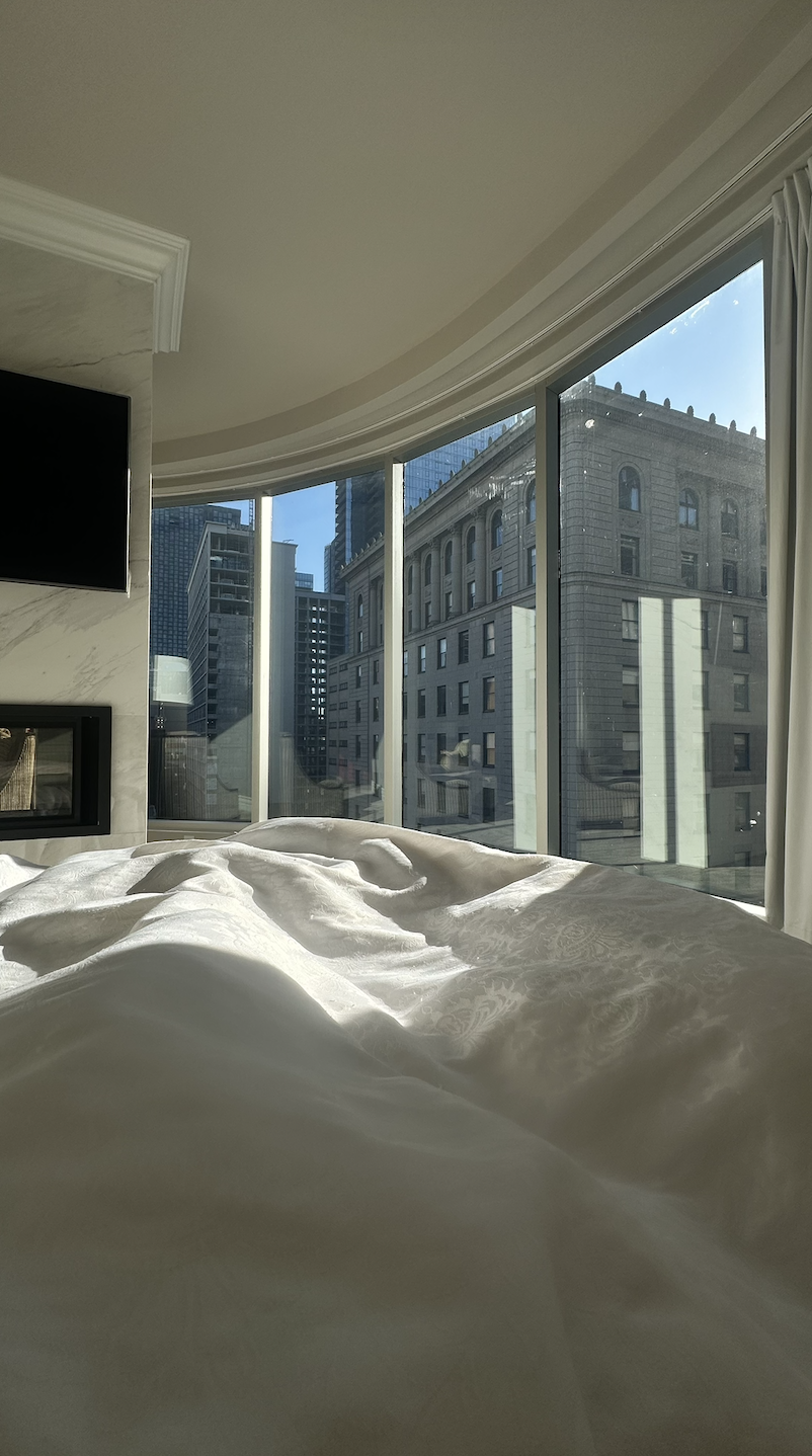 Morning sunlight over white hotel linens beside curved windows overlooking historic downtown Montreal architecture at Hotel Birks