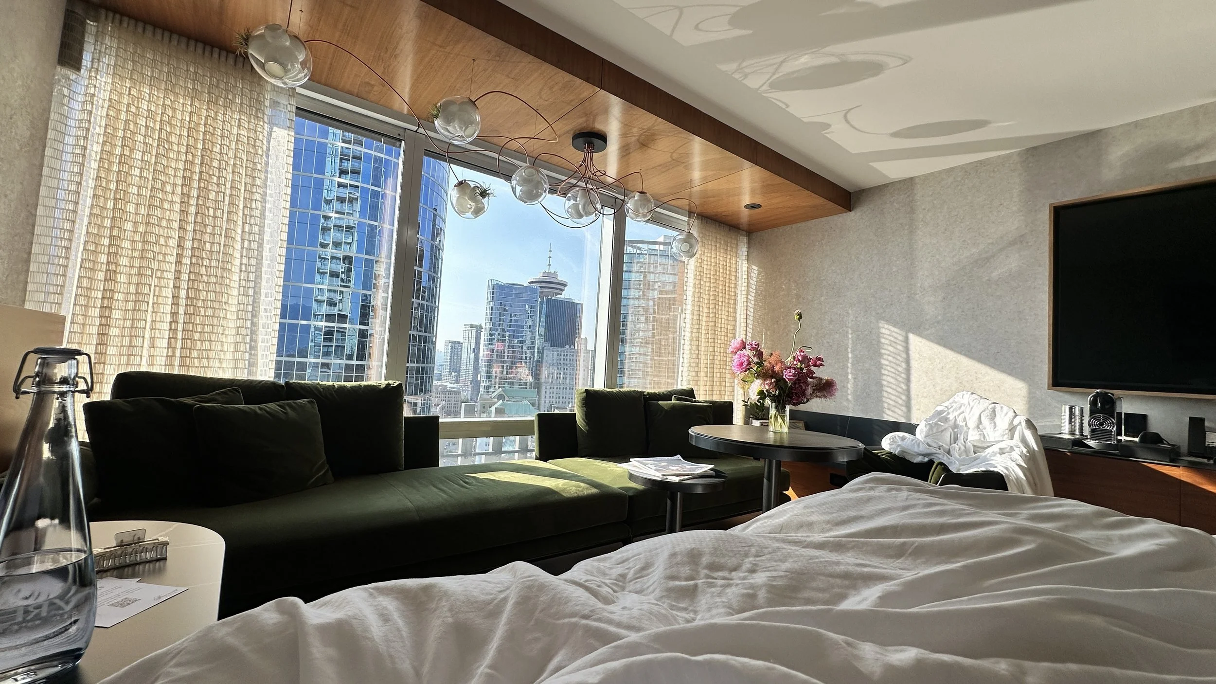 Sunlit luxury suite at Fairmont Pacific Rim in Vancouver, featuring green velvet sofas, modern lighting, fresh flowers and panoramic city views