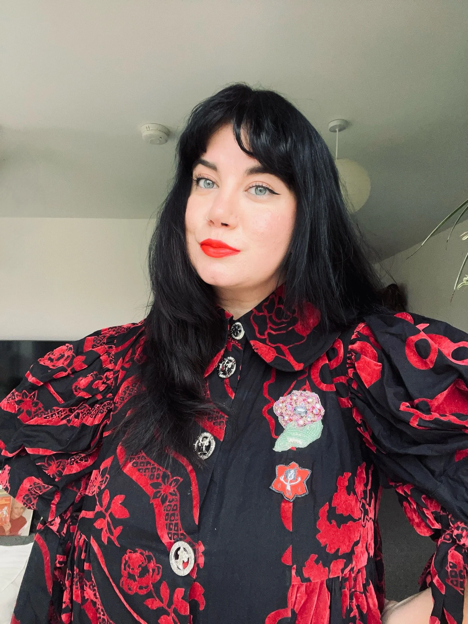 A woman with long black hair, blue eyes, and red lipstick, wearing a black and red patterned jacket with embroidered patches, standing indoors