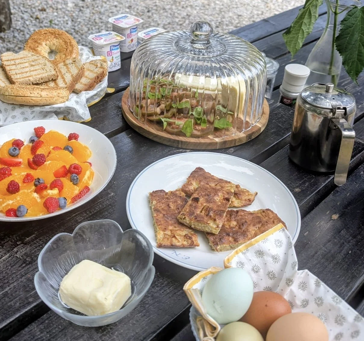 Frühstückstisch mit Obst, Eiern, Backwaren, Butter, Joghurt, Salat, und dekorativen Elementen auf einem Holztisch im Freien.