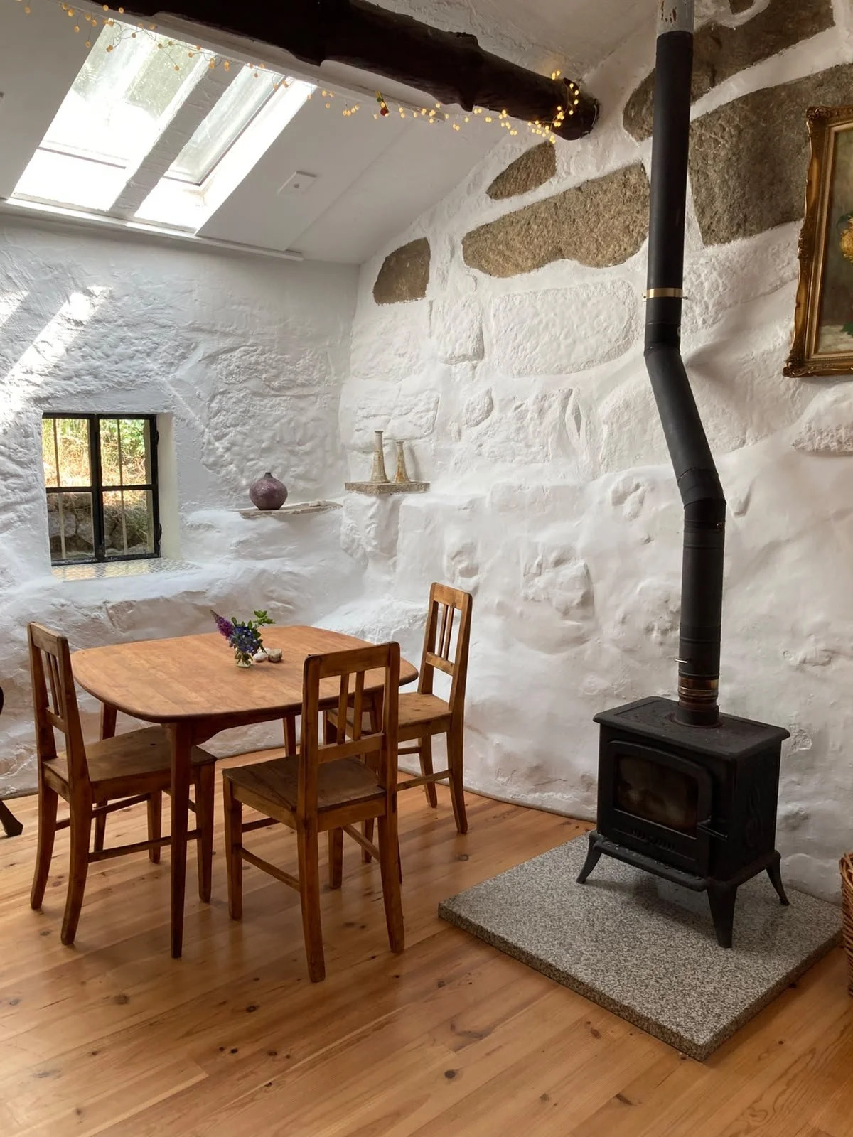 A cozy dining room with white textured stone walls and wooden floors. There is a wooden dining table with four matching chairs, a small vase with purple flowers on the table, a small window, and a skylight allowing natural light. A black wood-burning stove with a pipe extends upwards, and decorative items are displayed on wall shelves. A painting hangs on the wall.