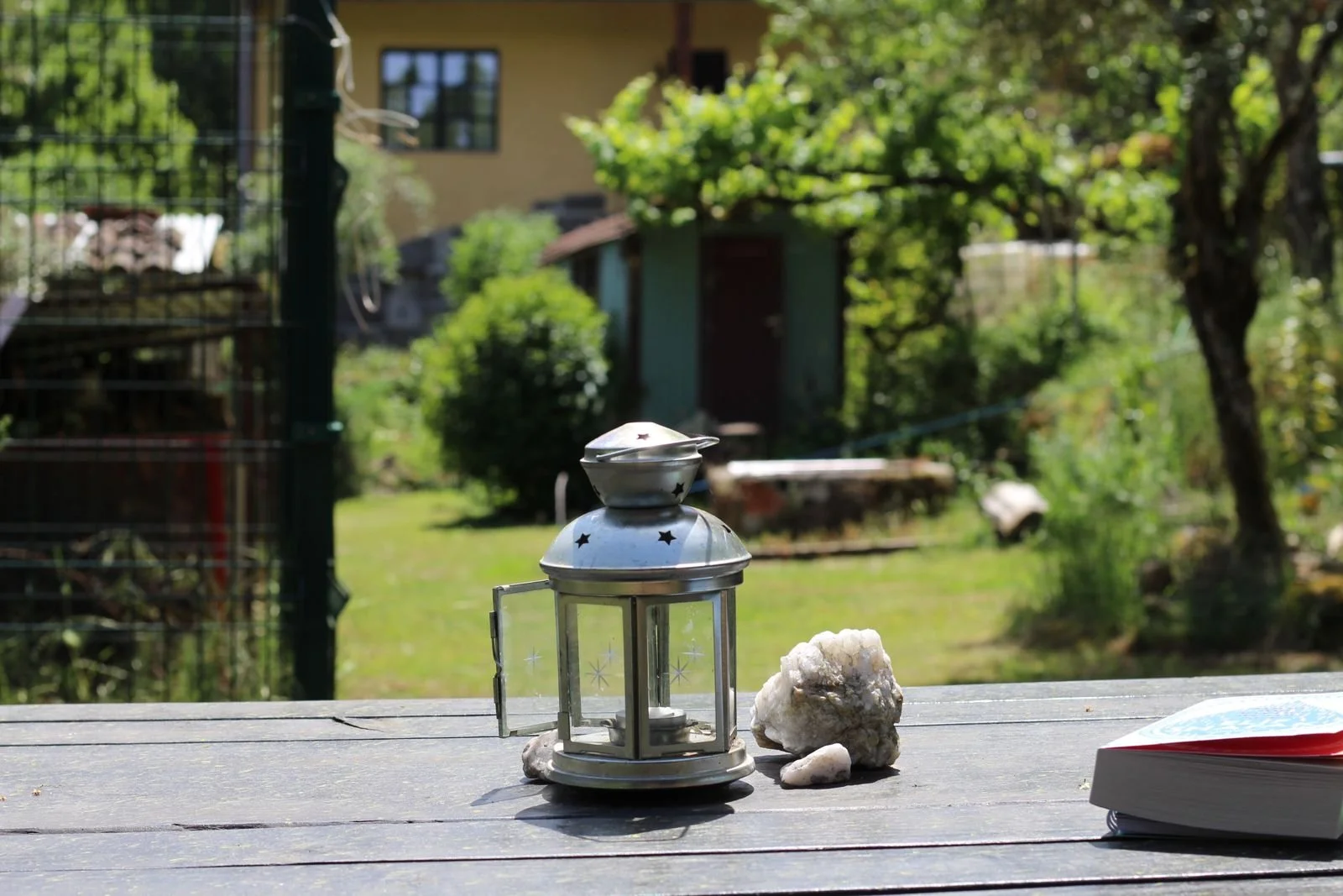 Silberne Laterne, weißer Stein, offene Buch auf einem Holztisch im Garten bei sonnigem Wetter.