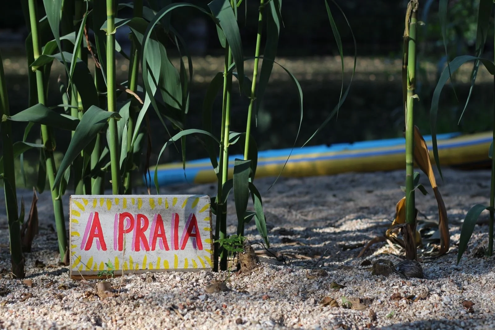 Boden mit Sand und Maispflanzen, kleines Handgemachtes Schild mit roten Buchstaben 'A PRAIA', im Hintergrund die Wasserrutsche auf einer Strand- oder Gartenanlage.