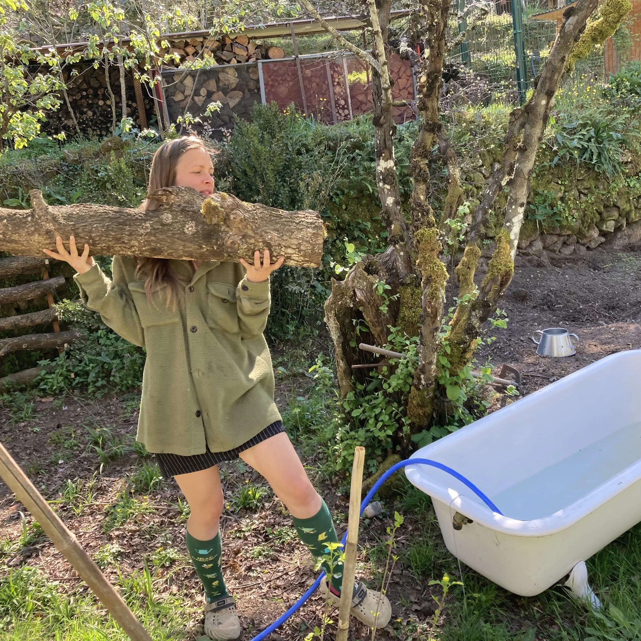 A girl wearing green rain boots, a green coat, and striped shorts is holding a large branch in a garden, standing near a white plastic tub connected to a blue hose, with trees, shrubs, and neatly stacked firewood in the background.