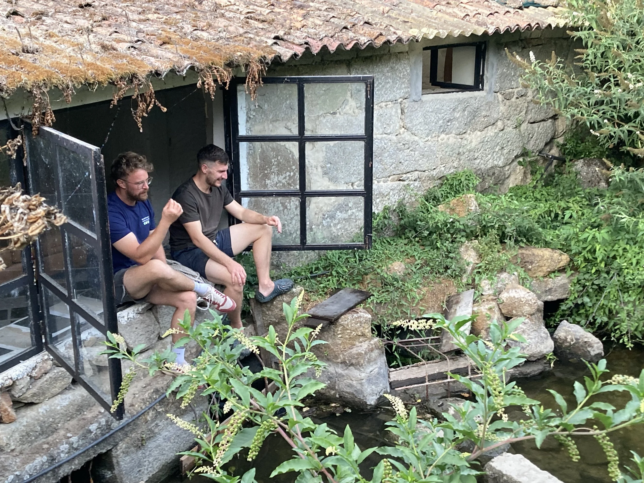 Two men sitting on rocks by a small pond outside a stone house with small windows and a tiled roof, surrounded by greenery.