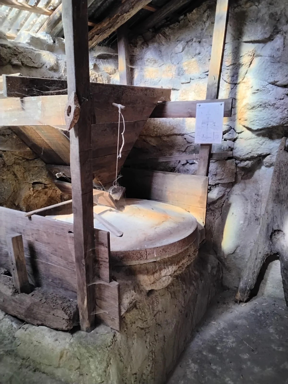 An old watermill wheel made of wood and stone, with a wooden cover and stone wall in the background, inside an underground or basement space.